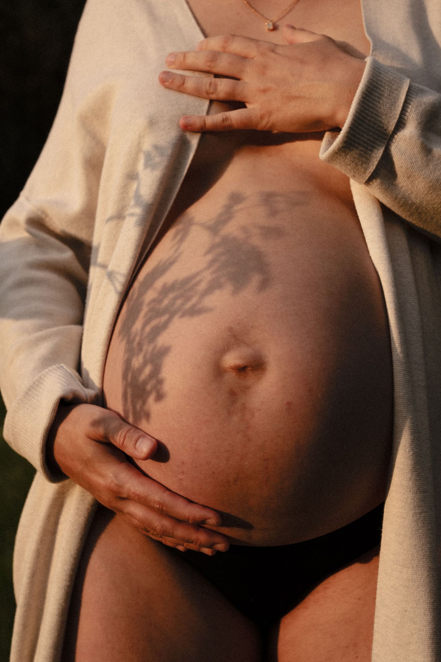 Pregnant woman outdoors holding baby bump with floral shadow pattern cast across bare belly in warm light. by Soft Sacred Studios