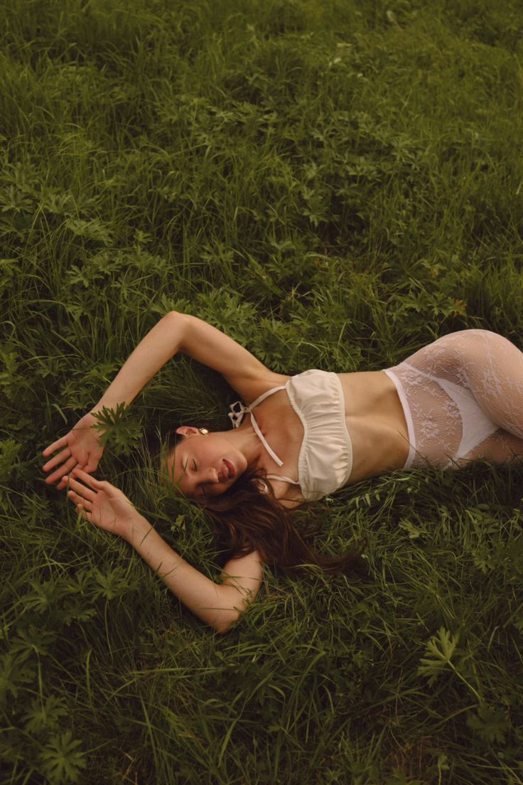 Young woman in white lace lingerie lying peacefully in lush green grass outdoors. By Soft Sacred Studios.