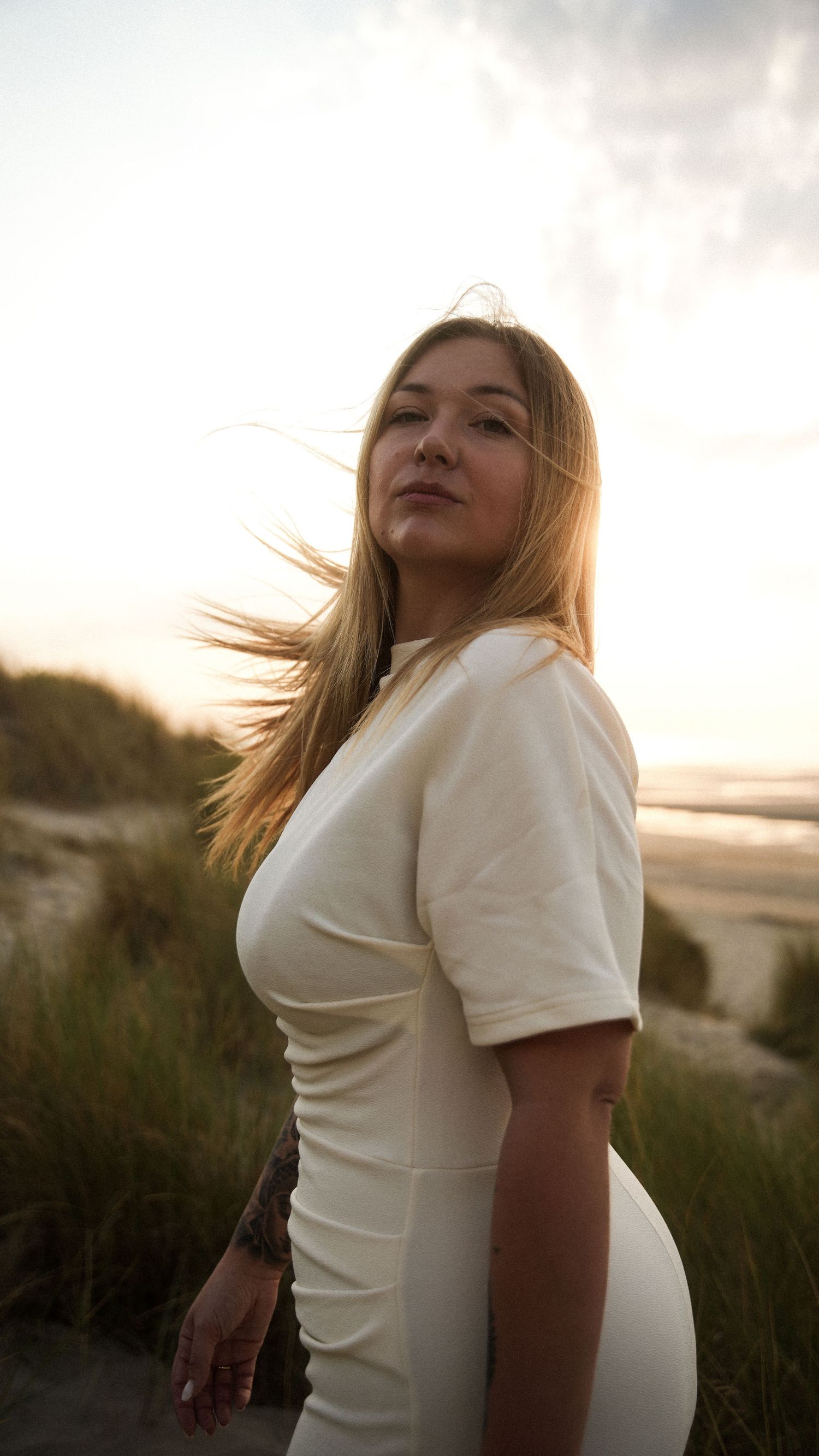 Blonde woman in white dress standing on beach dunes at sunset with hair blowing. By Soft Sacred Studios.