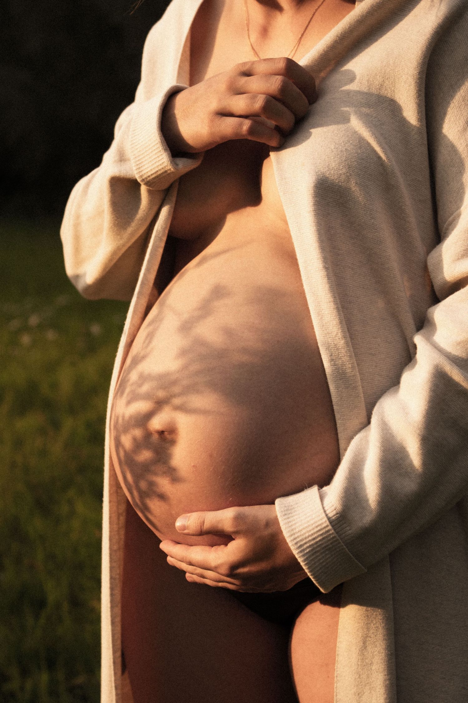Pregnant woman in beige cardigan holding bare belly with soft tree shadow in warm golden sunlight. by Soft Sacred Studios