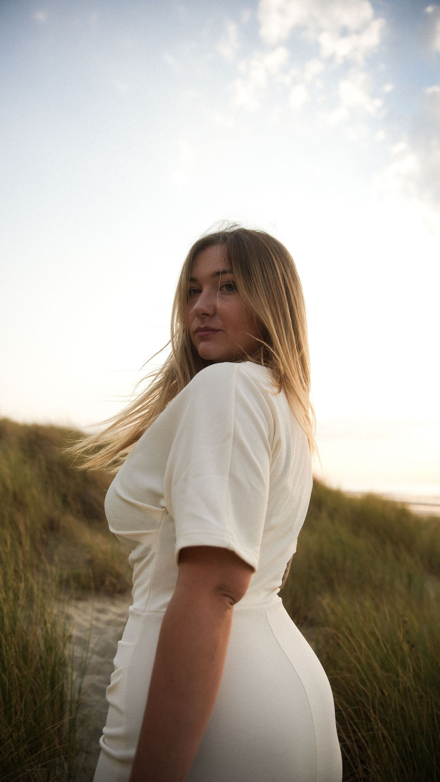 Blonde woman in white dress stands on sandy beach dunes at golden sunset hour. By Soft Sacred Studios.