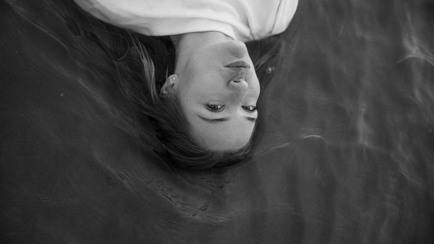 Overhead black and white portrait of woman floating in dark water, looking up. by Soft Sacred Studios