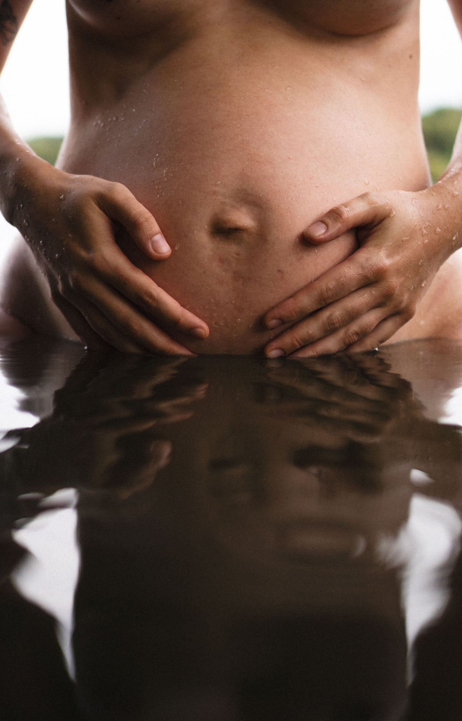 Close-up of pregnant belly held by two hands at water level, with a mirror-like reflection on the surface. By Soft Sacred Studios.