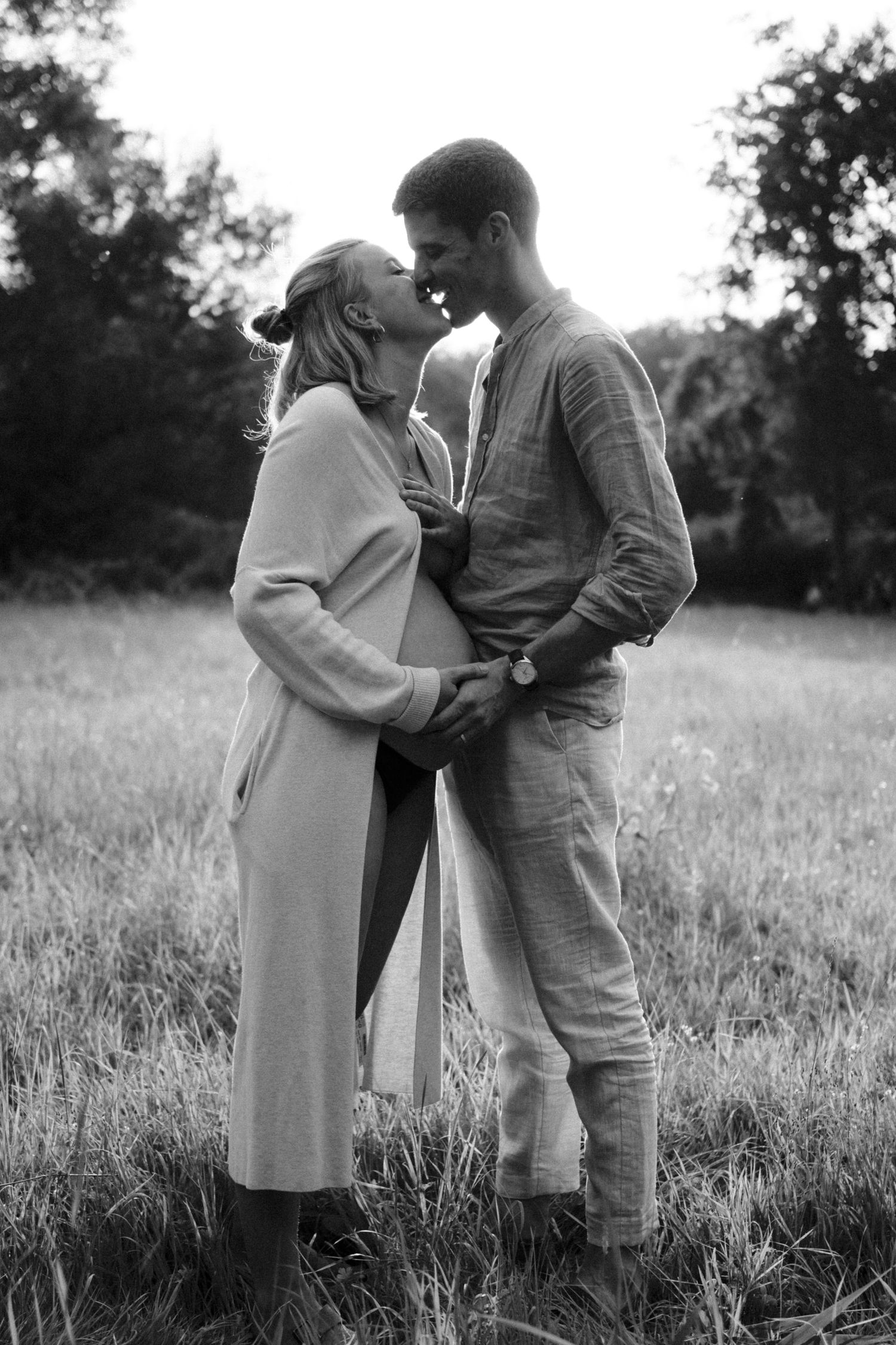 Black and white photo of couple sharing a tender kiss in a grassy field during maternity shoot. By Soft Sacred Studios.
