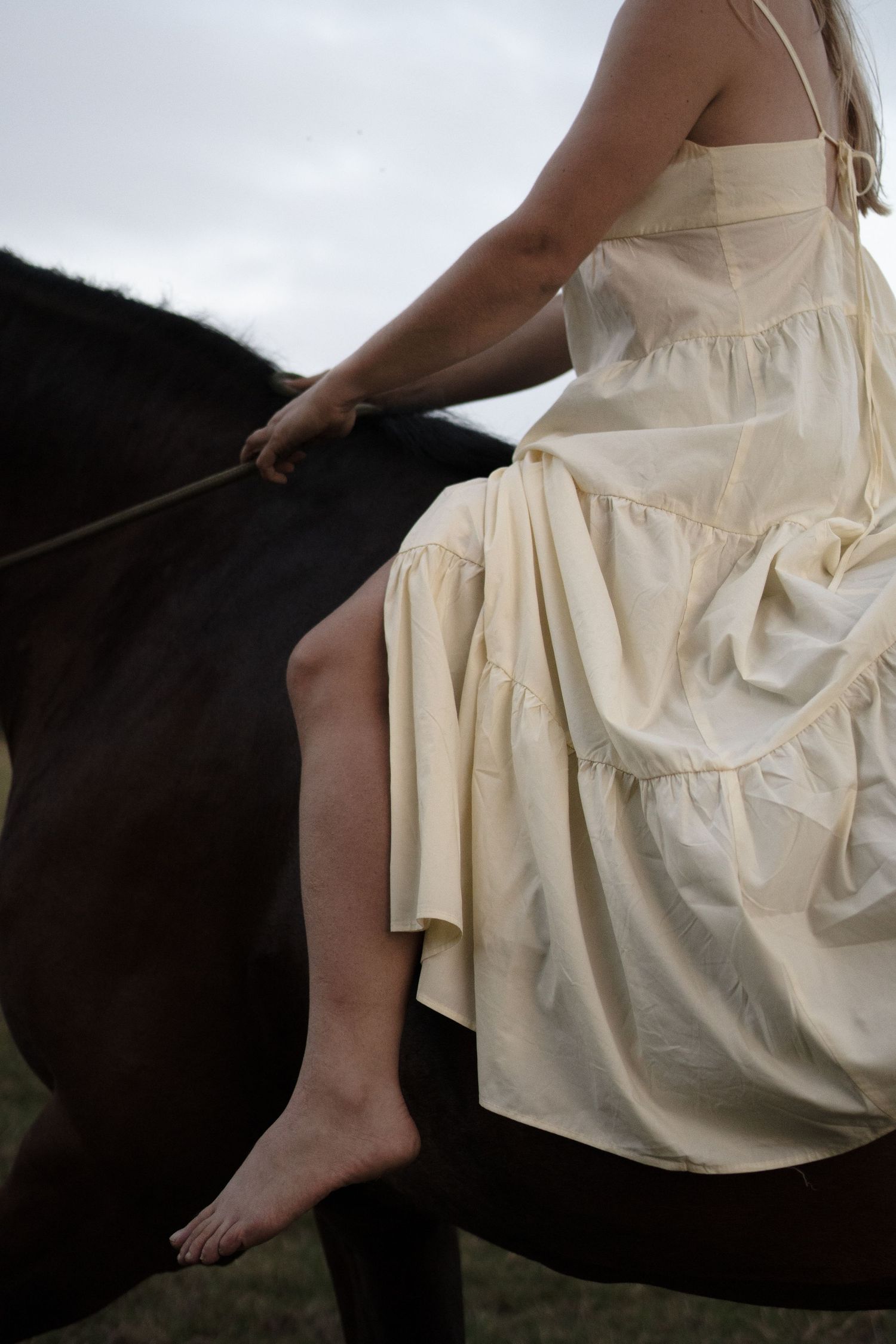 Close-up of a barefoot woman in cream dress riding bareback on a dark horse outdoors. by Soft Sacred Studios