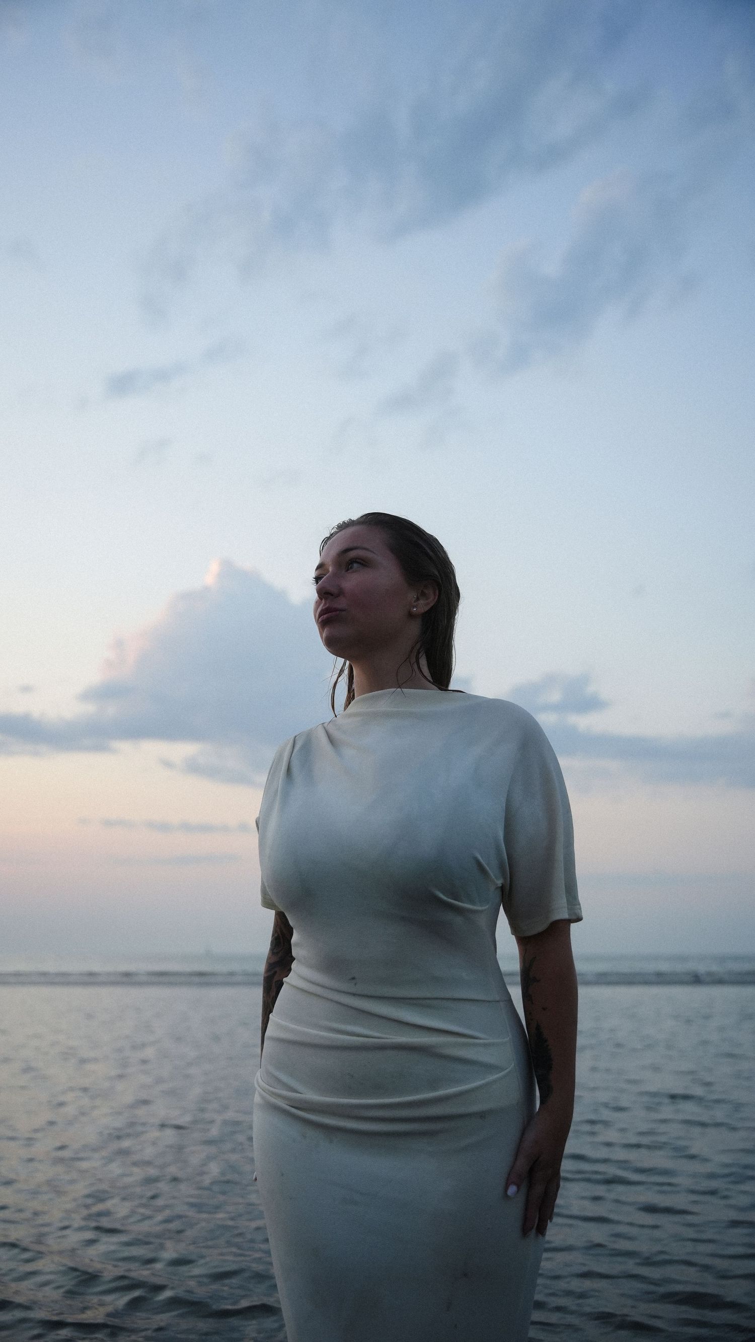 Woman in white dress standing on beach at sunset, looking upward against a dramatic cloudy sky. by Soft Sacred Studios