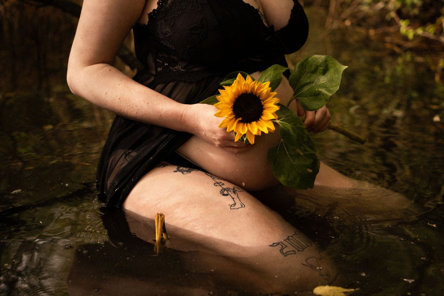 Tattooed pregnant woman in black lace dress holding sunflower while sitting in shallow forest stream. By Soft Sacred Studios.