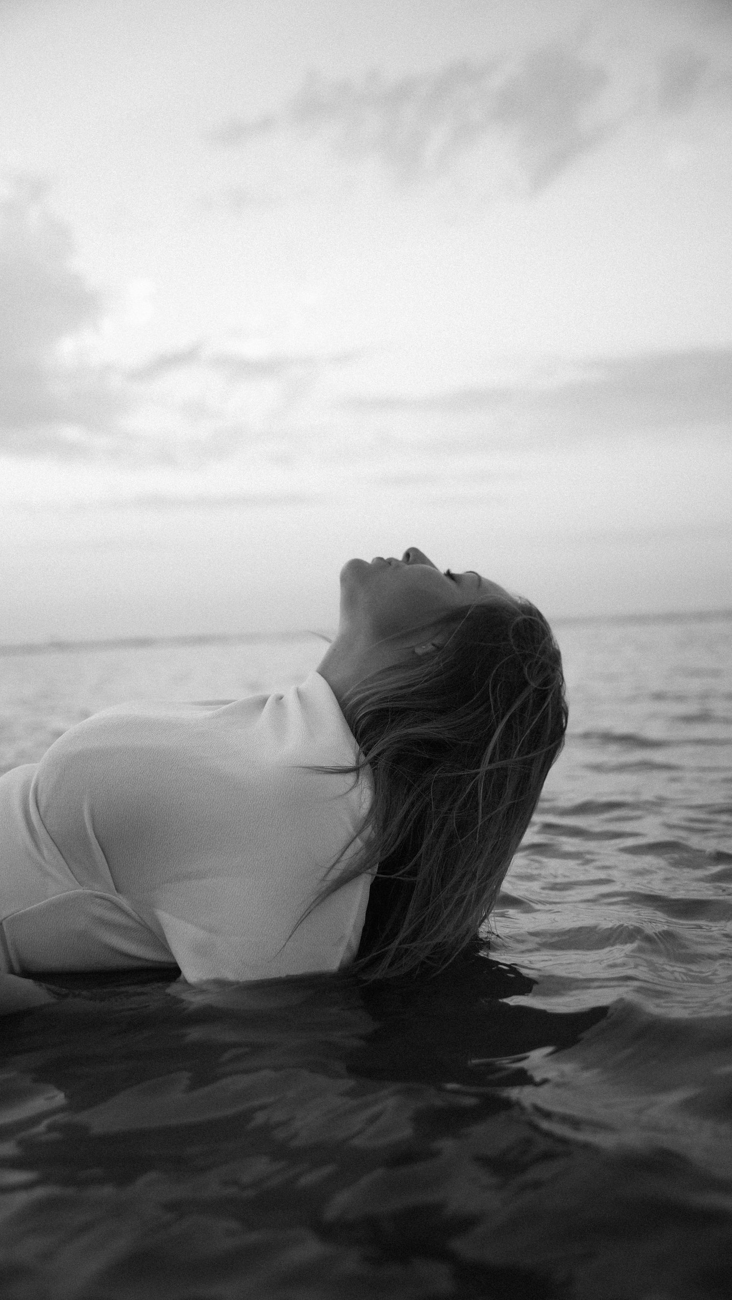 Woman in white shirt emerging from ocean waves with head back, dramatic black and white photo. by Soft Sacred Studios