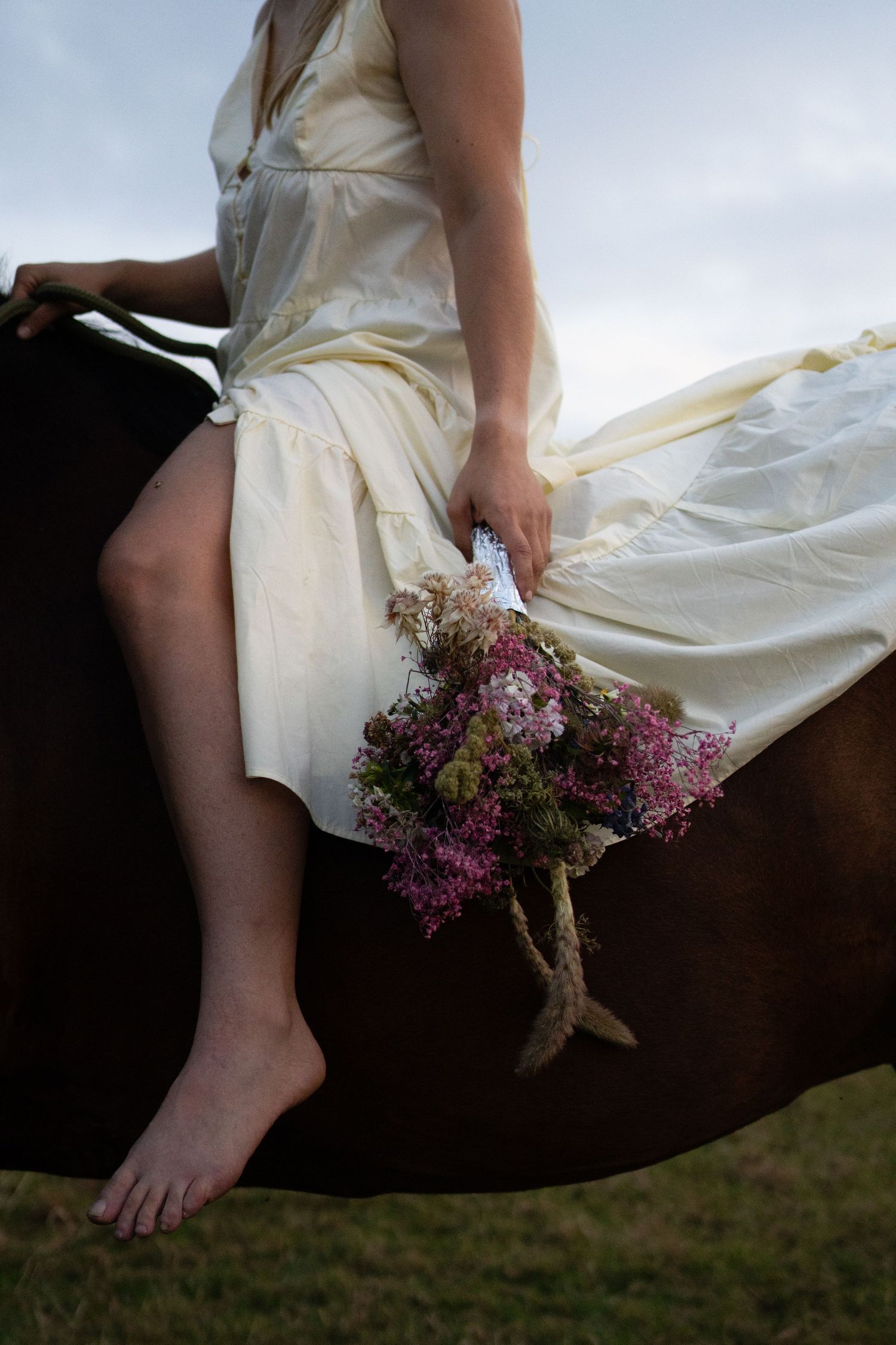Young woman in cream dress sitting bareback on a dark horse in a meadow. By Soft Sacred Studios.