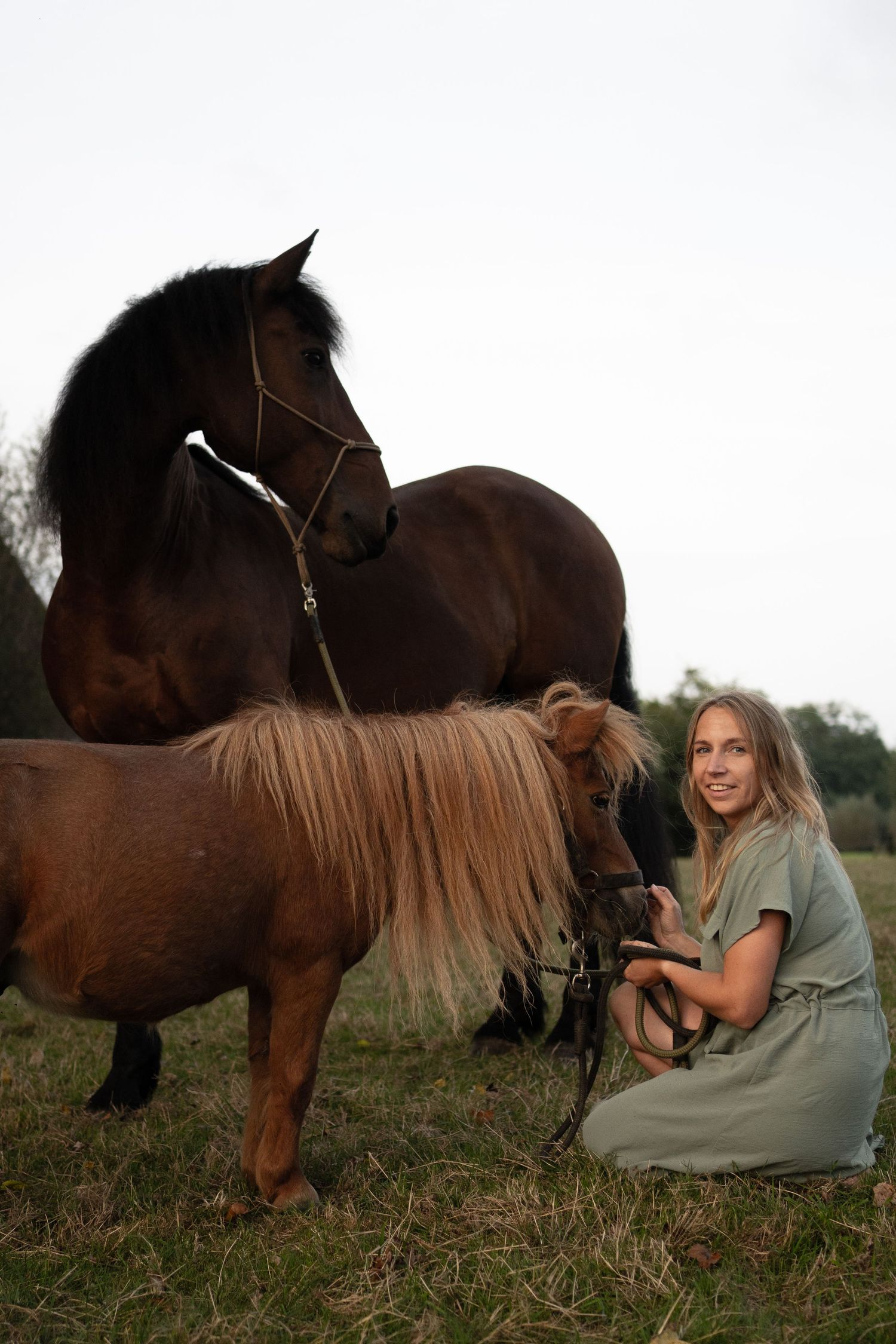 Smiling blonde woman in green dress crouching beside a pony and large dark horse in a field. by Soft Sacred Studios