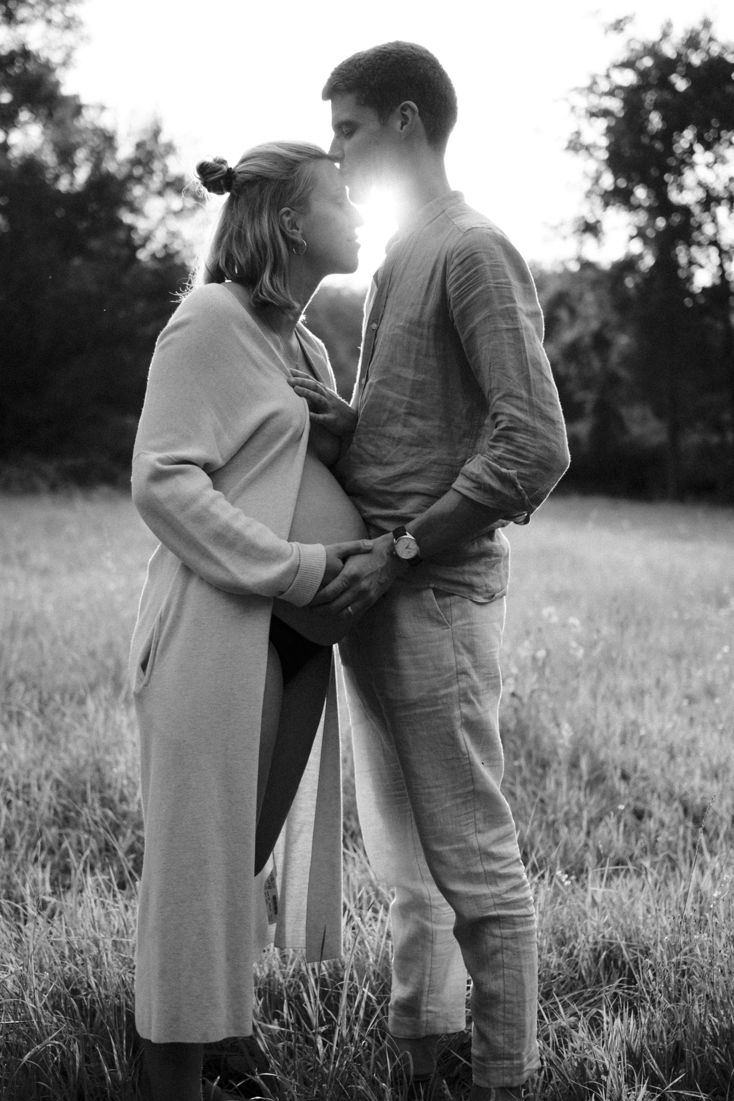 Backlit black and white maternity photo of couple embracing in a sunlit field at golden hour. By Soft Sacred Studios.