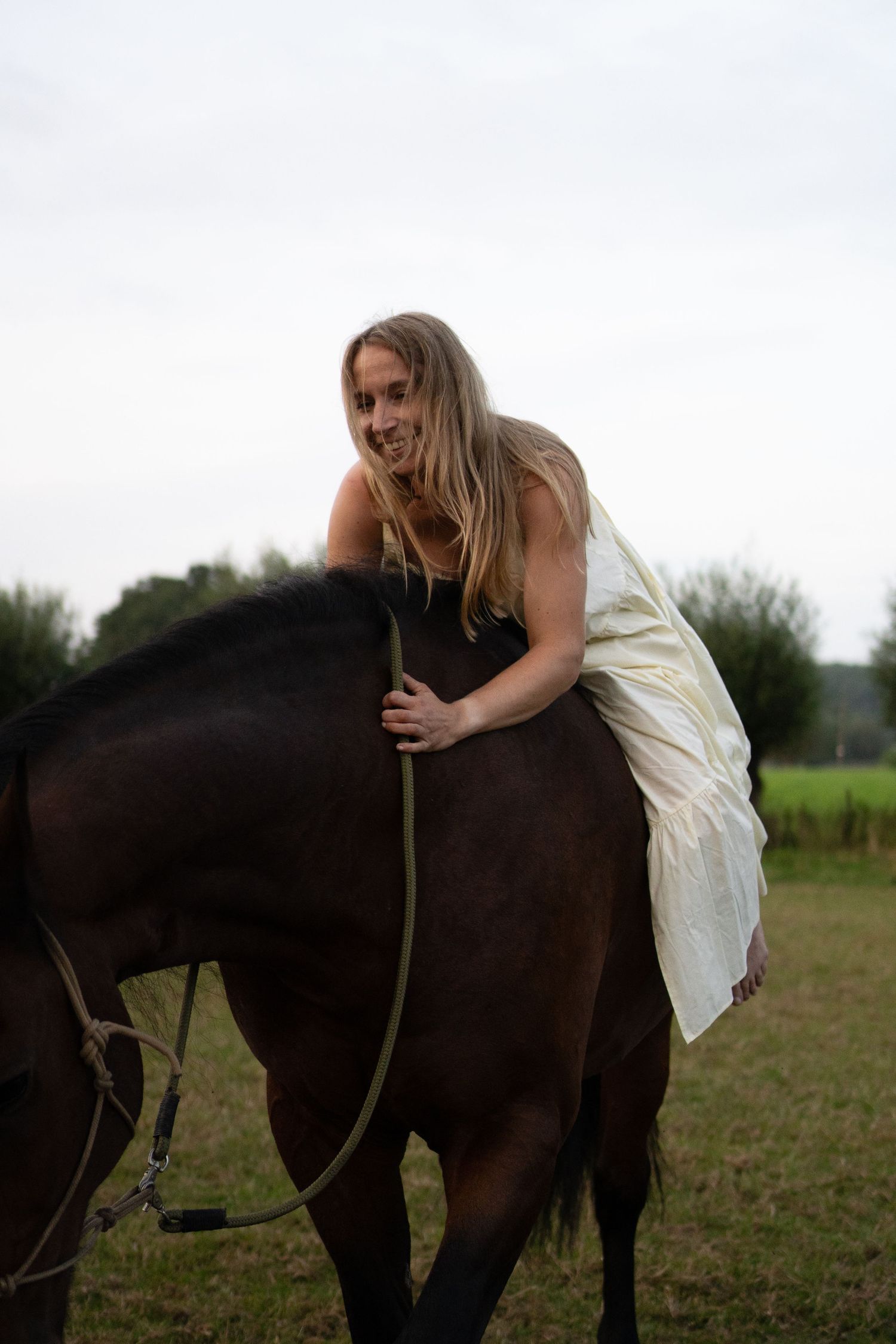 Young woman in cream dress sitting bareback on a dark horse in a meadow. By Soft Sacred Studios.