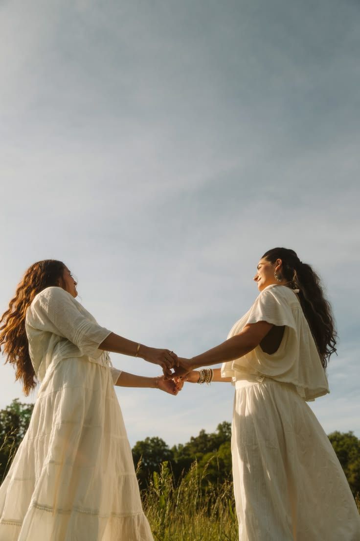 Two women in white dresses holding hands and spinning together in a sunny field. By Soft Sacred Studios.