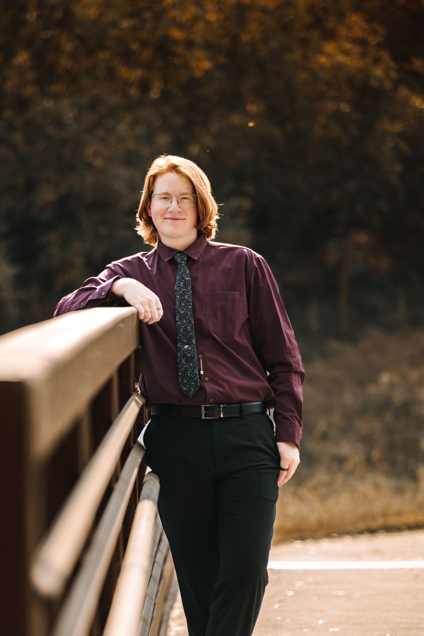 young man posin for senior pictures wearin a marron shirt. posing on a bridge for his senior pictures.