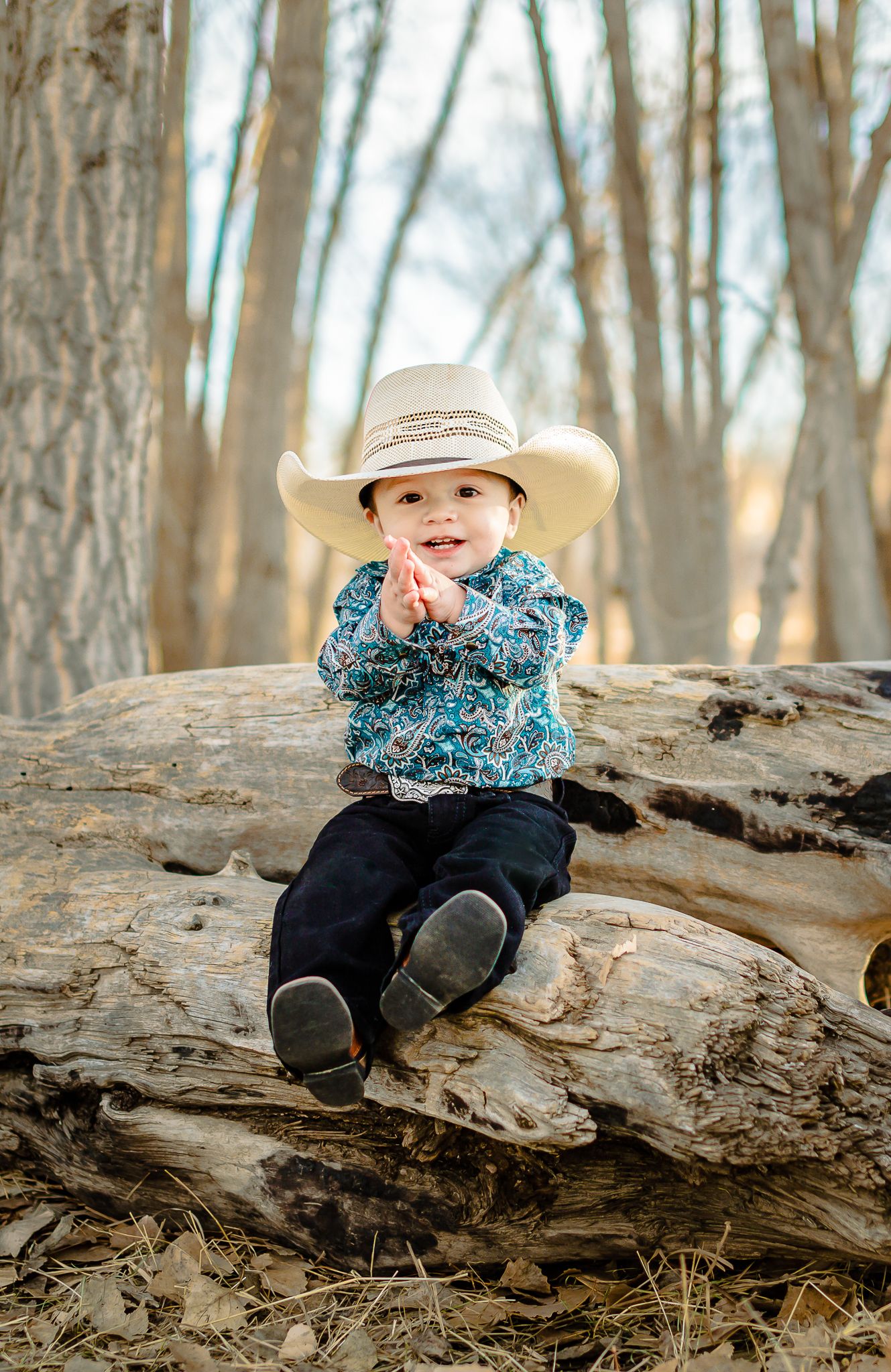 one year old boy smiling while the greeley photographer takes his pictures while he smiles in this western style outfit. sitting on a log