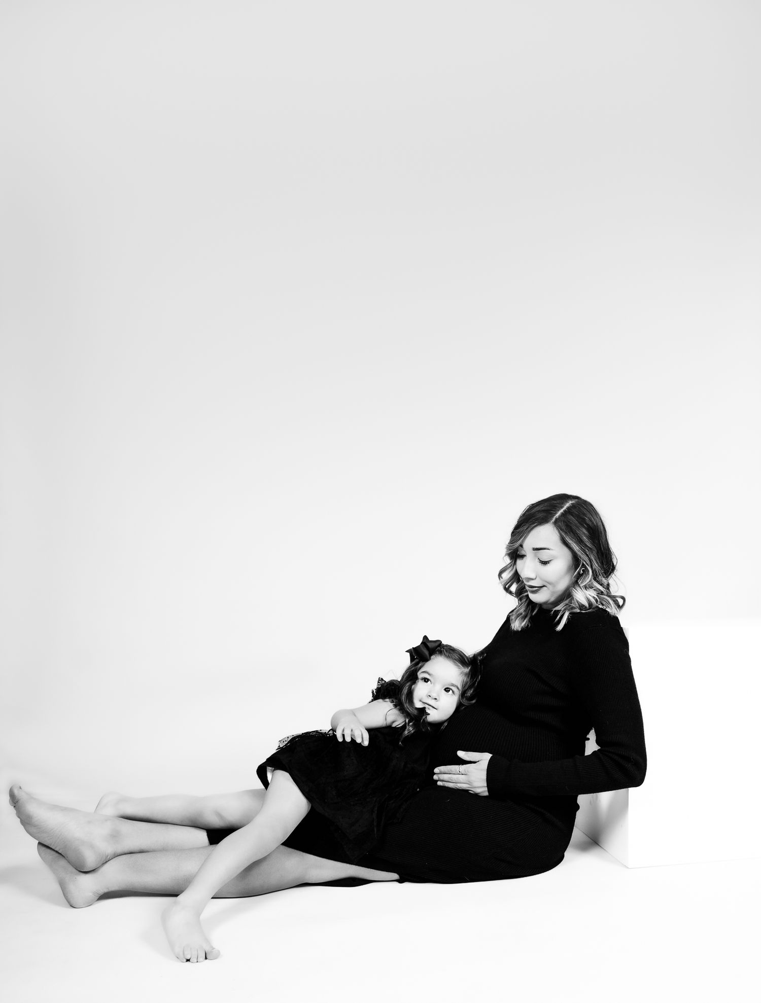 mom and daughter leaning on a posing block wearing black dresses. the mom is looking down and the child is looking at the camera posing for