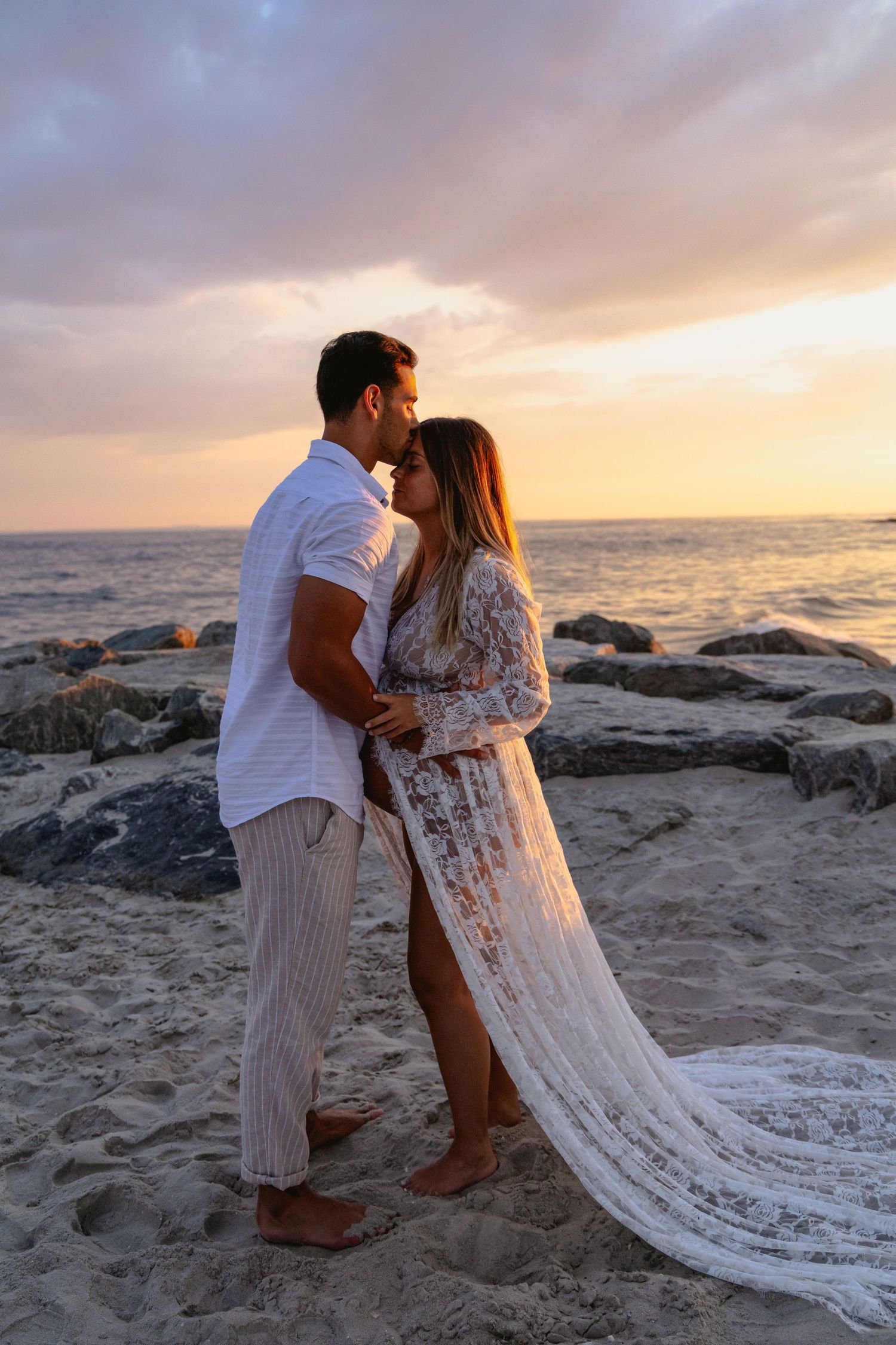 Romantic couple embracing at sunset on rocky beach, woman wearing flowing white lace maternity gown.