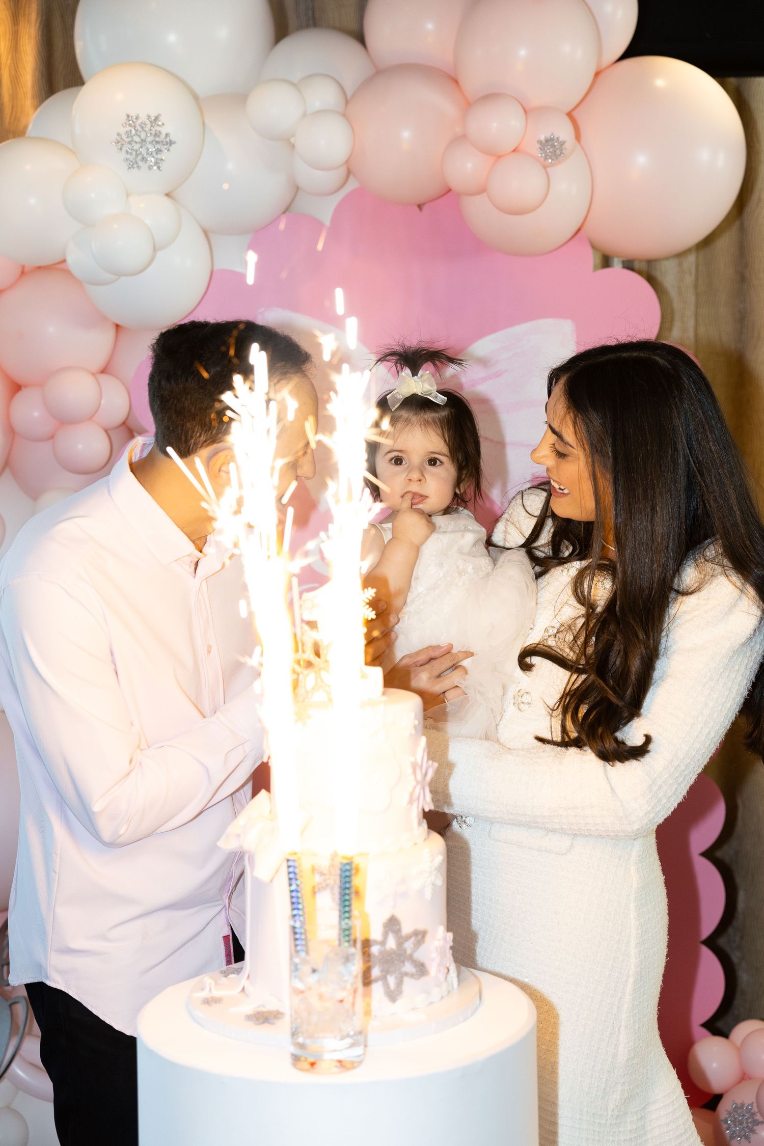 Parents with toddler girl celebrating first birthday with sparkling cake and pink balloon arch.