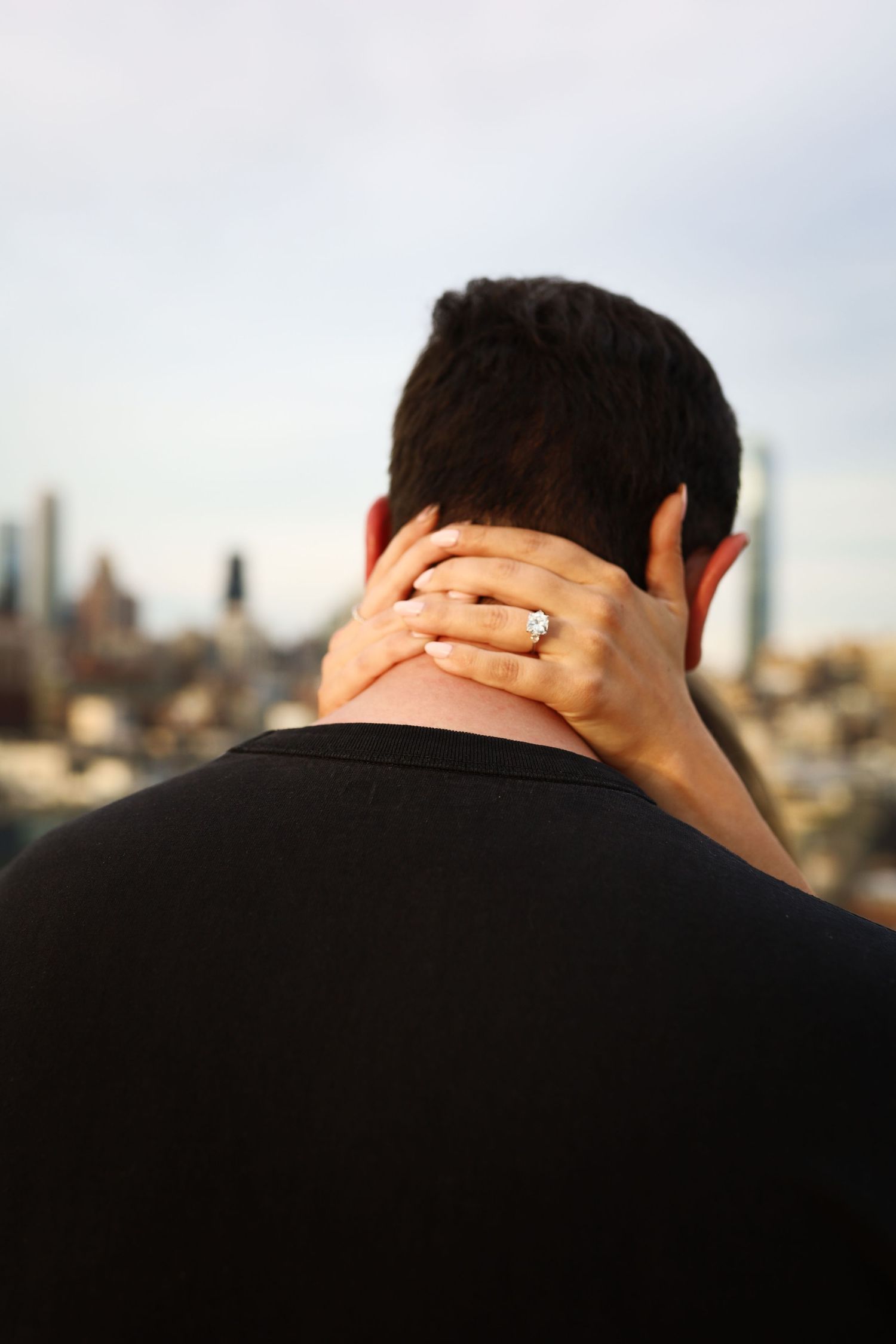 Woman resting hand with diamond engagement ring on mans neck with city skyline in background.