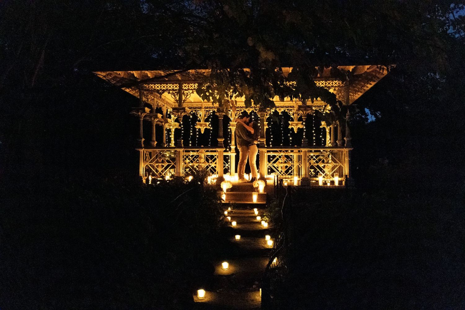 Romantic nighttime proposal at illuminated gazebo with candle-lined pathway in a dark garden setting.