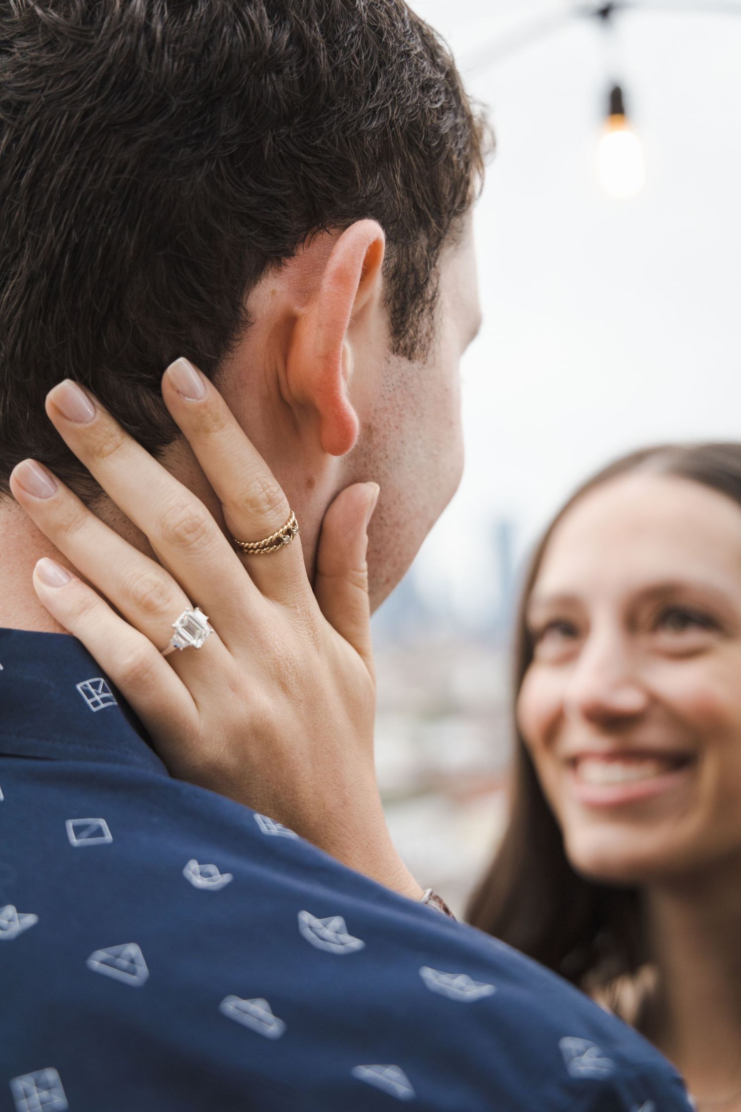 Close-up of smiling woman showing diamond engagement ring while embracing partner outdoors on rooftop.