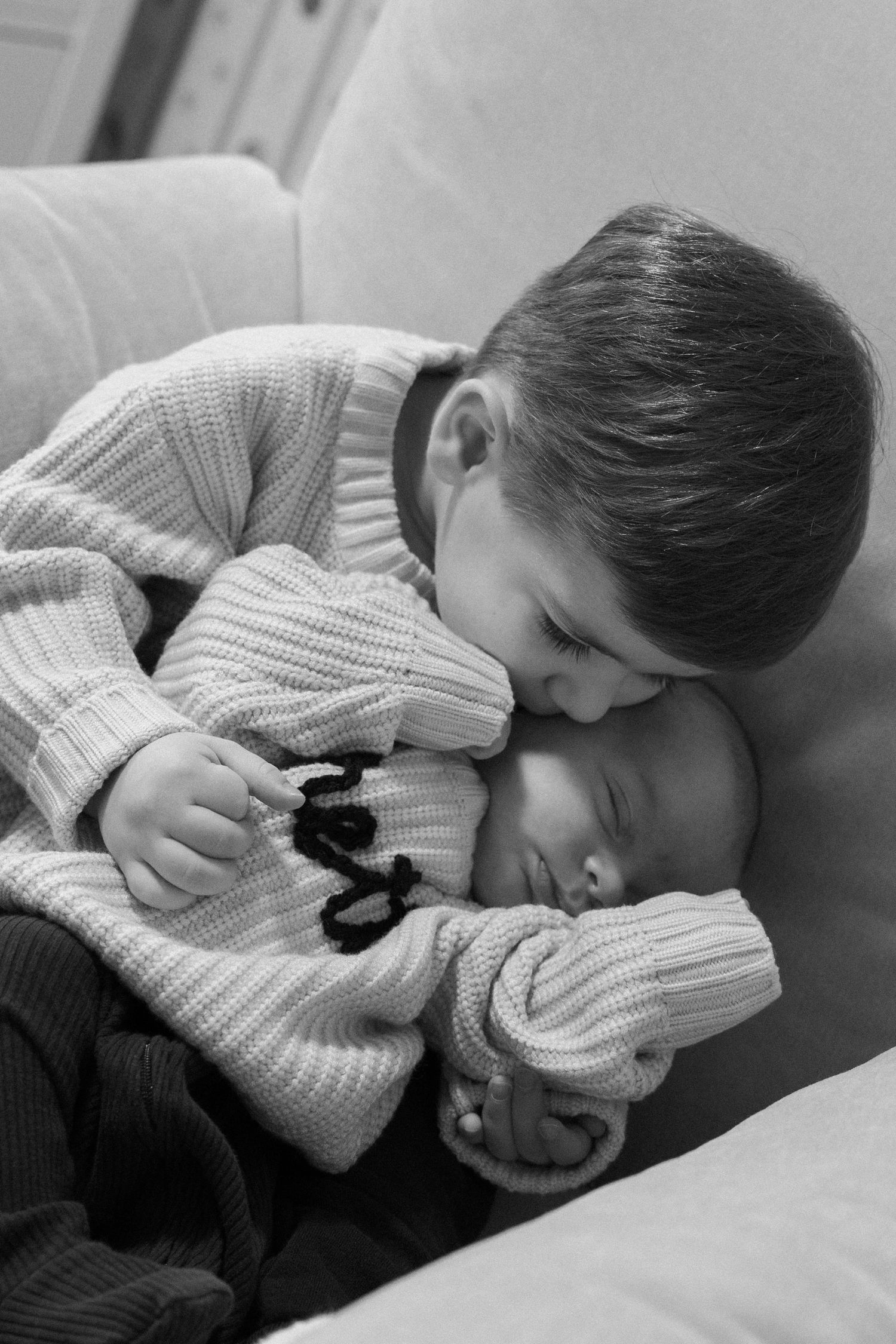 Black and white photo of young boy tenderly kissing sleeping newborn baby sibling while seated on a couch.