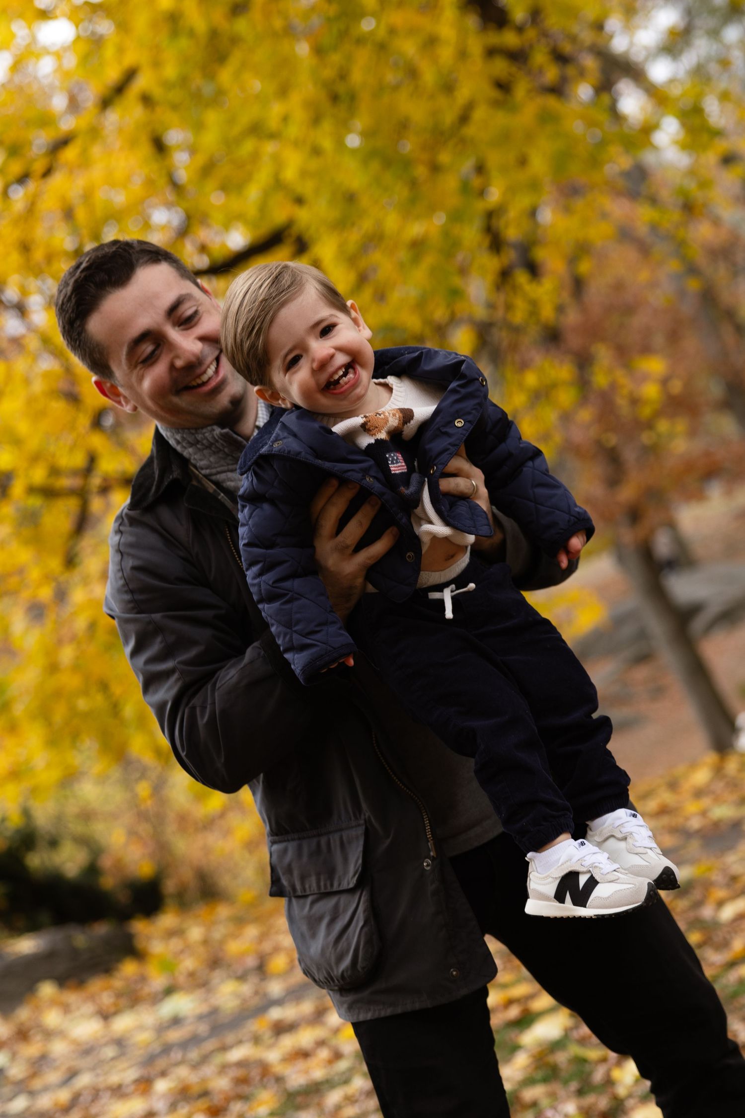 Father holding laughing toddler in autumn park with golden yellow foliage in background.