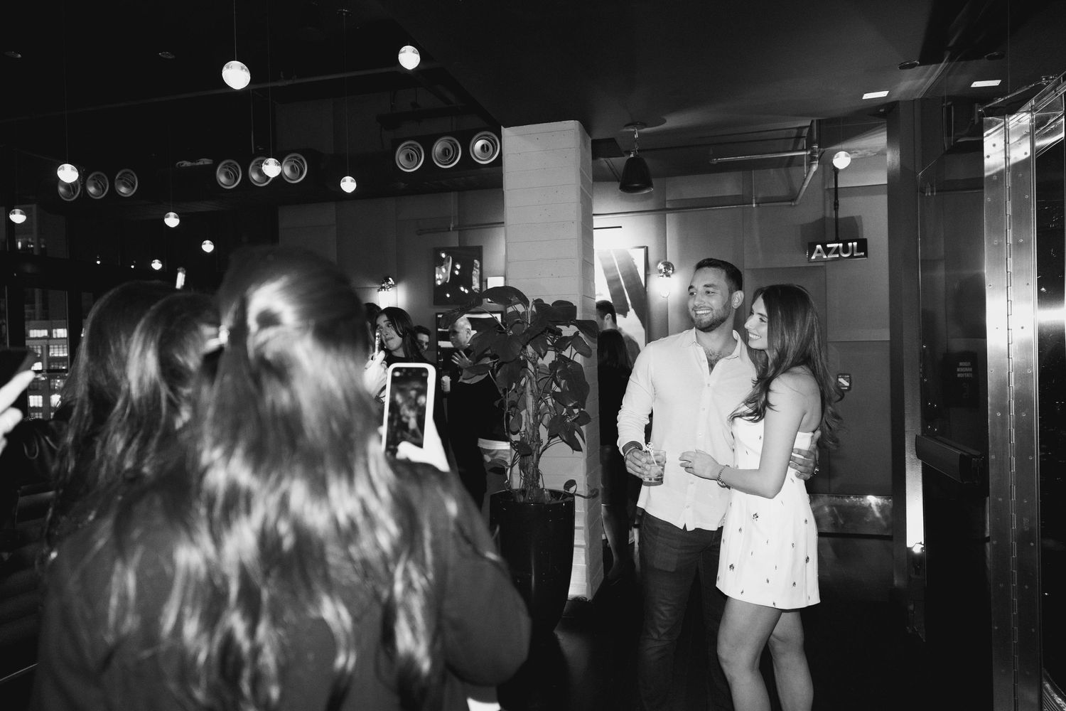 Black and white photo of a couple dancing at a party while guests take photos with smartphones.