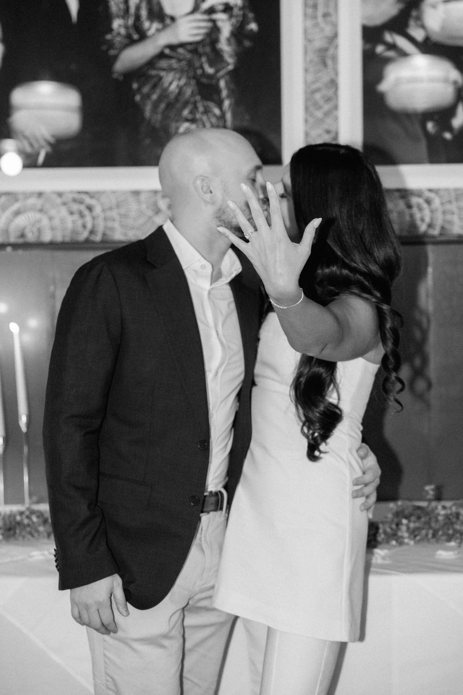 Black and white photo of couple kissing as bride shows off engagement ring to camera indoors.