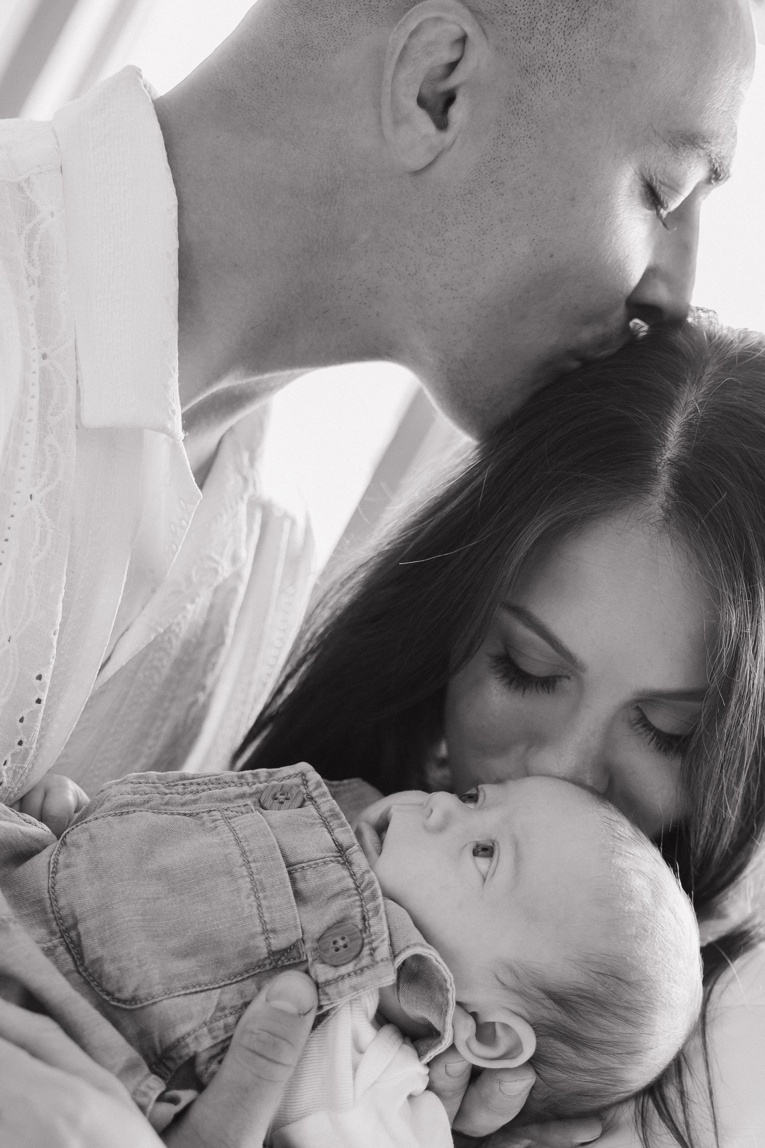 Parents tenderly kissing their newborn baby in a black and white portrait.