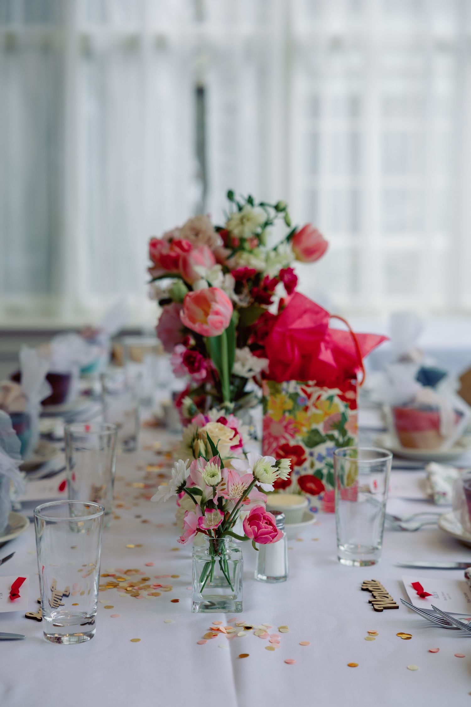 Festive table arrangement with pink and white floral centerpieces, gold confetti, and glassware for a celebratory event.