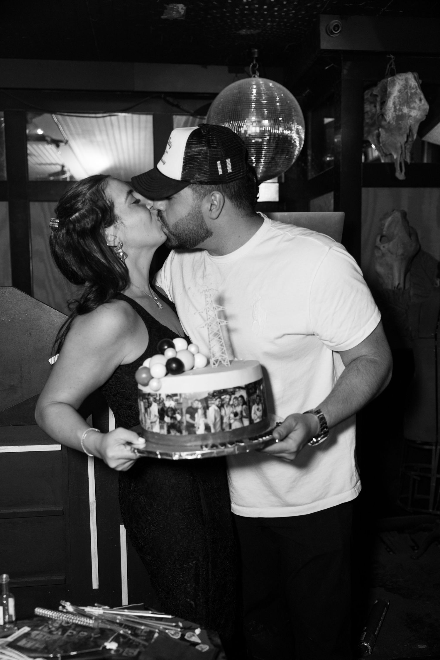 Black and white photo of a couple sharing a kiss while holding a decorated celebration cake.