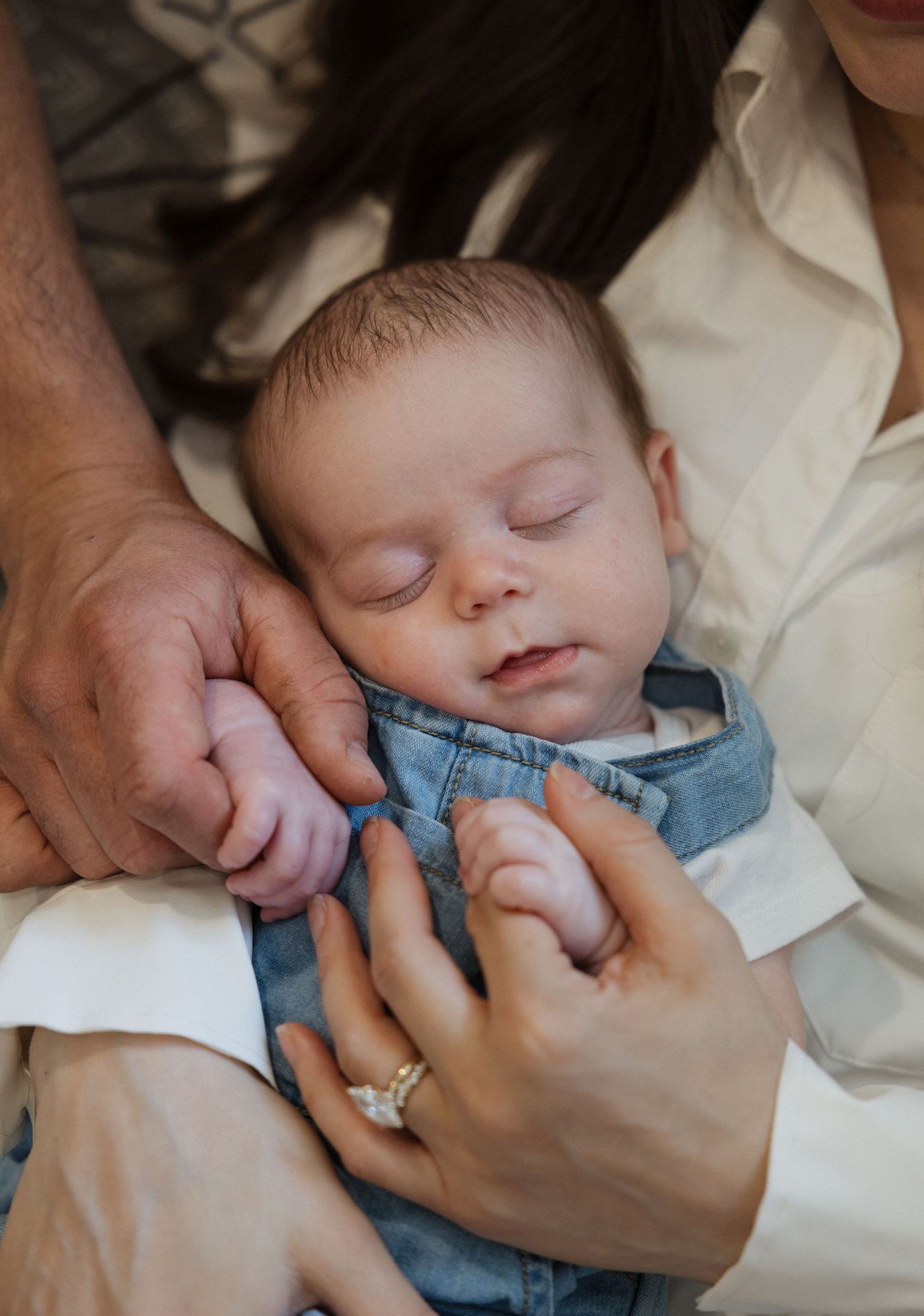 Sleeping newborn baby in denim overalls cradled in parents arms with mother wearing engagement ring.
