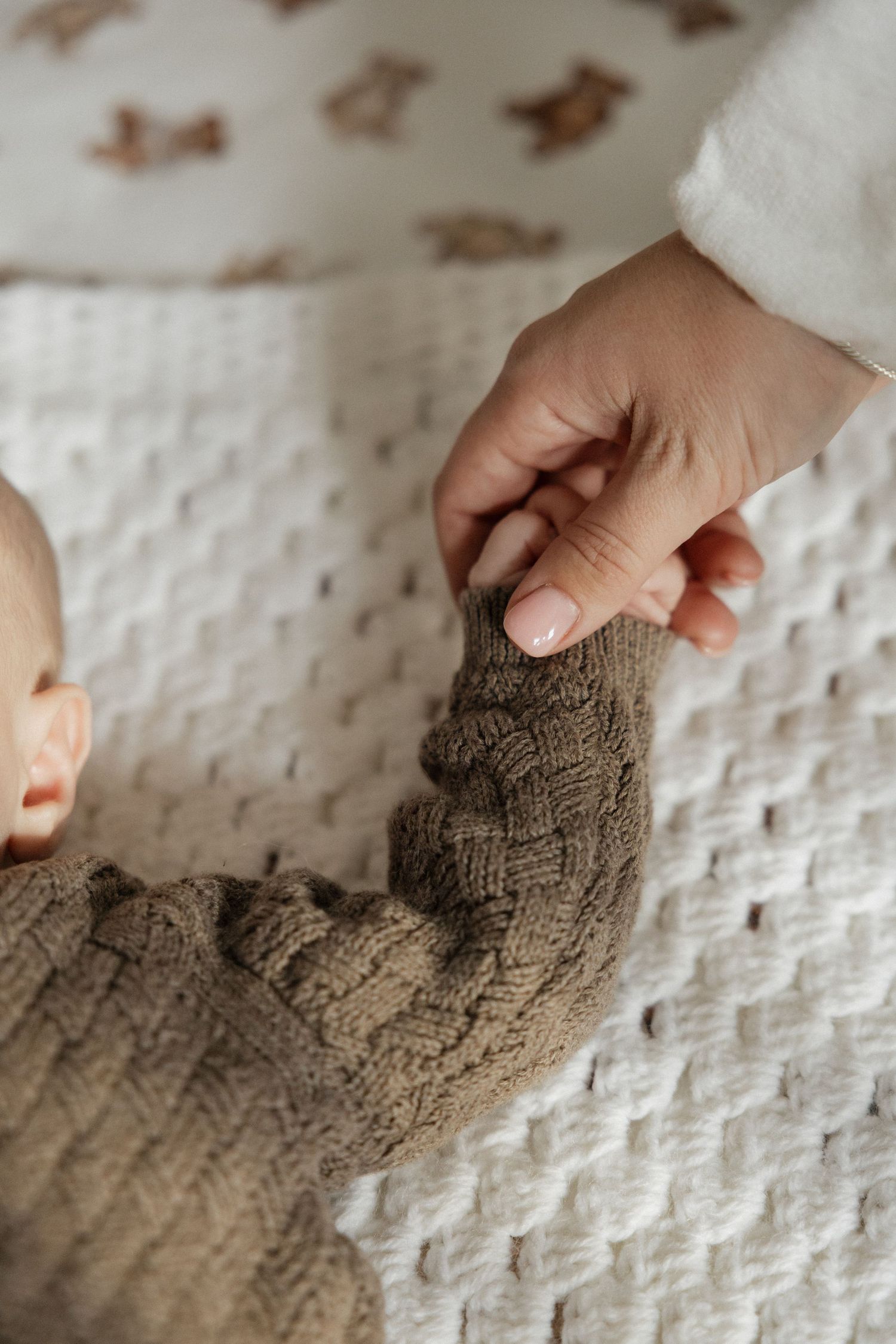 Parent gently holding newborn babys hand dressed in brown knit sweater on white textured blanket.