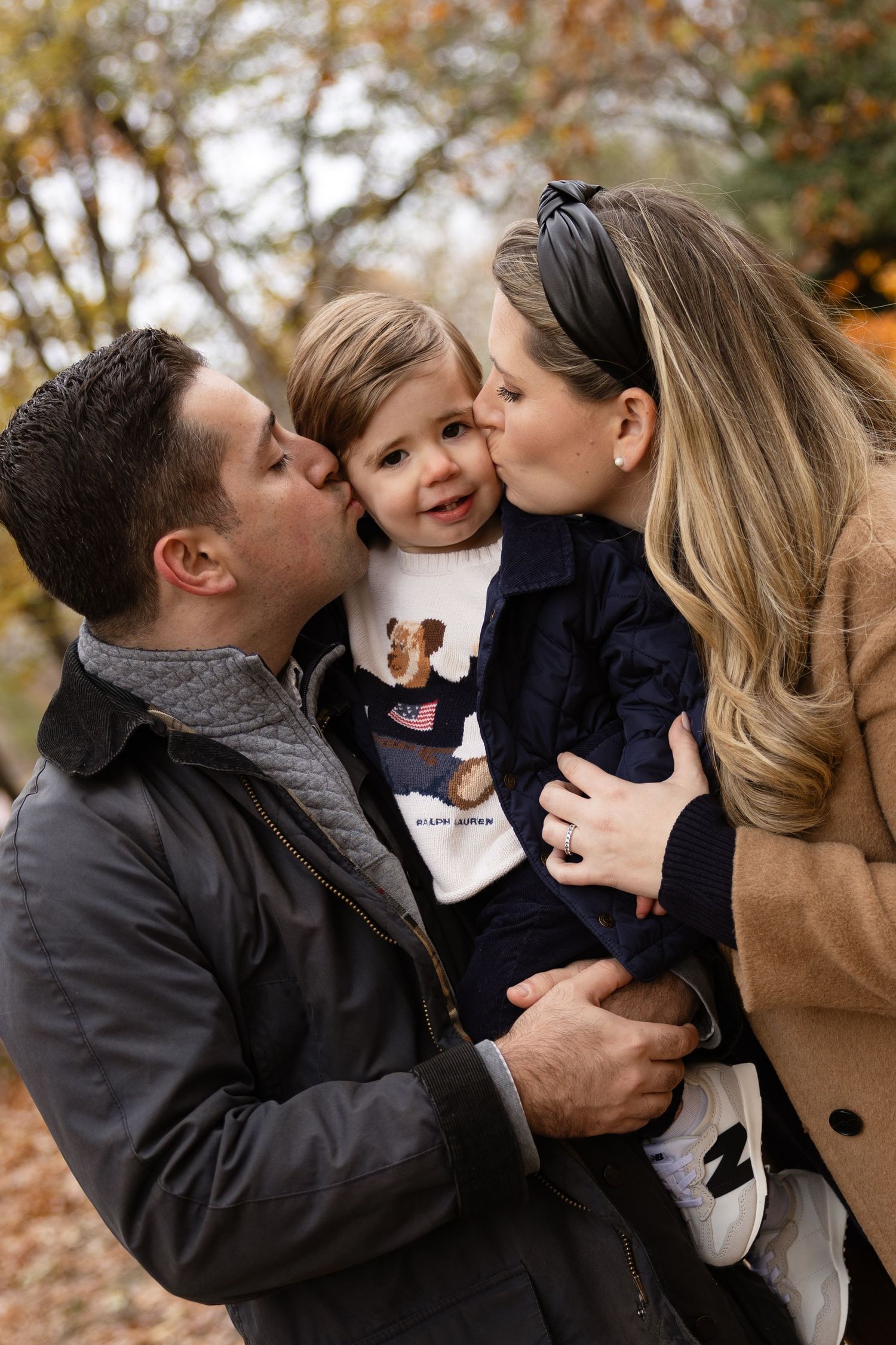 Family portrait with parents kissing toddler on cheeks outdoors in autumn park with colorful fall foliage.