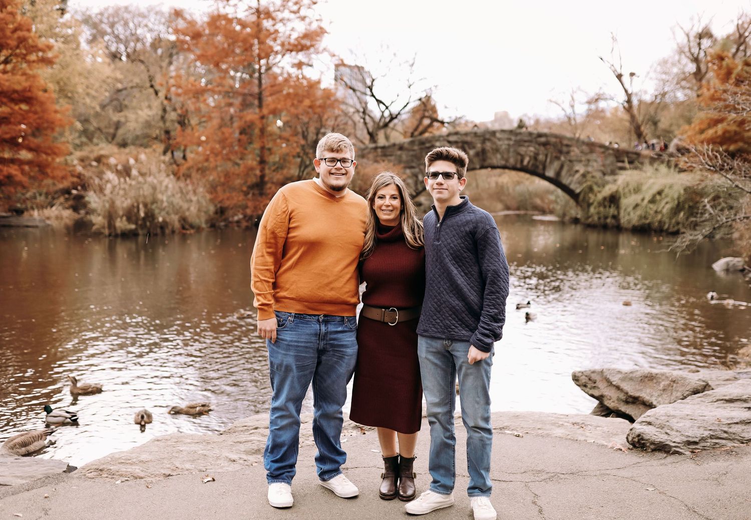 Family of three poses together by pond with stone bridge amid autumn foliage in park.