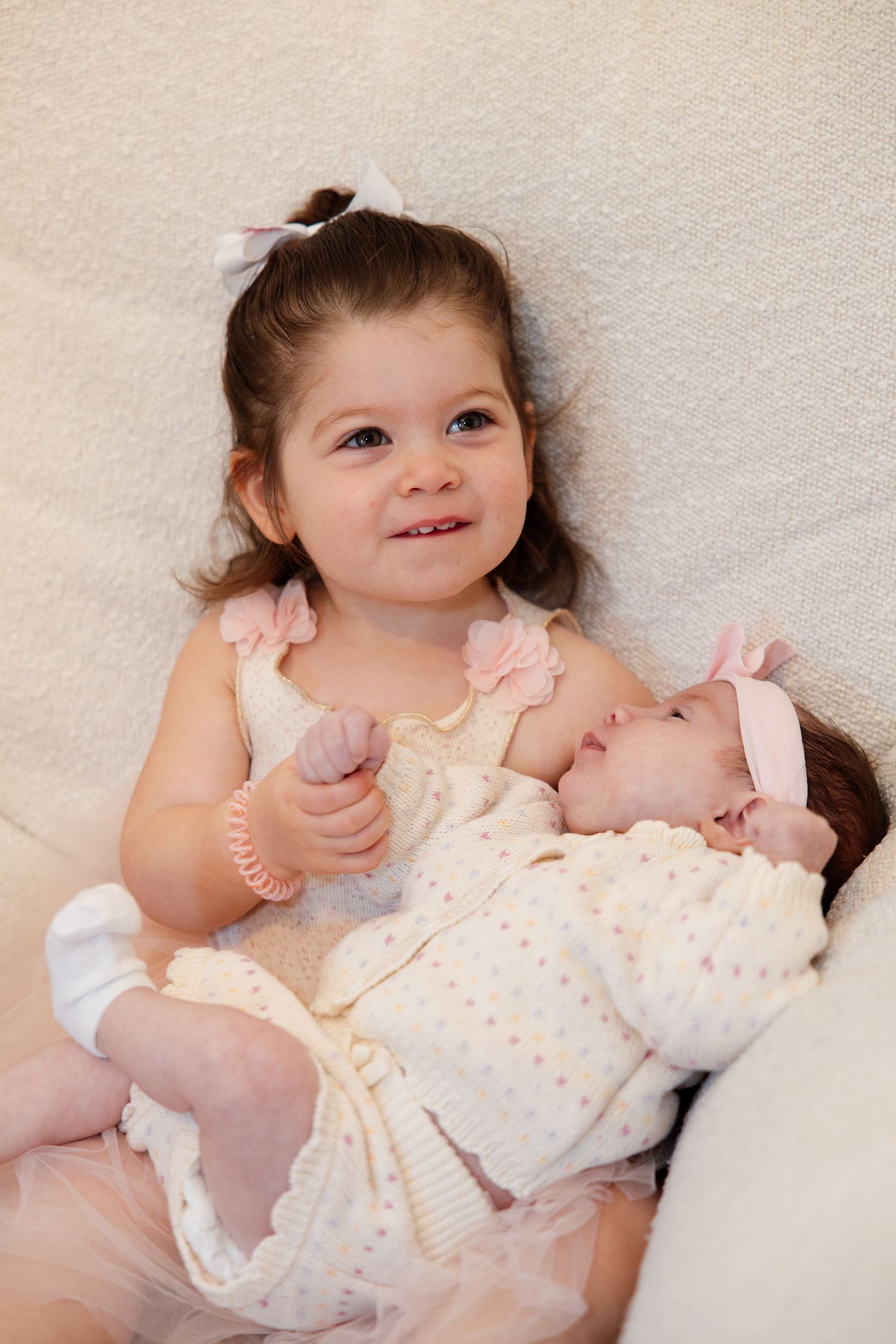 Smiling toddler girl in pink dress gently holding sleeping newborn baby sister in cream knit outfit.