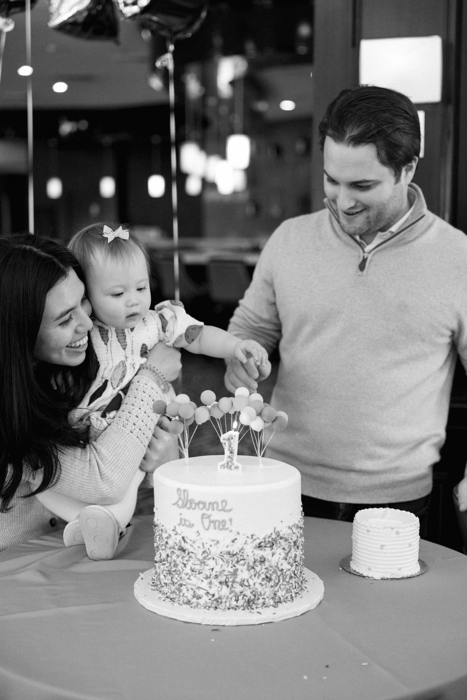 Black and white photo of parents helping baby touch first birthday cake at party.