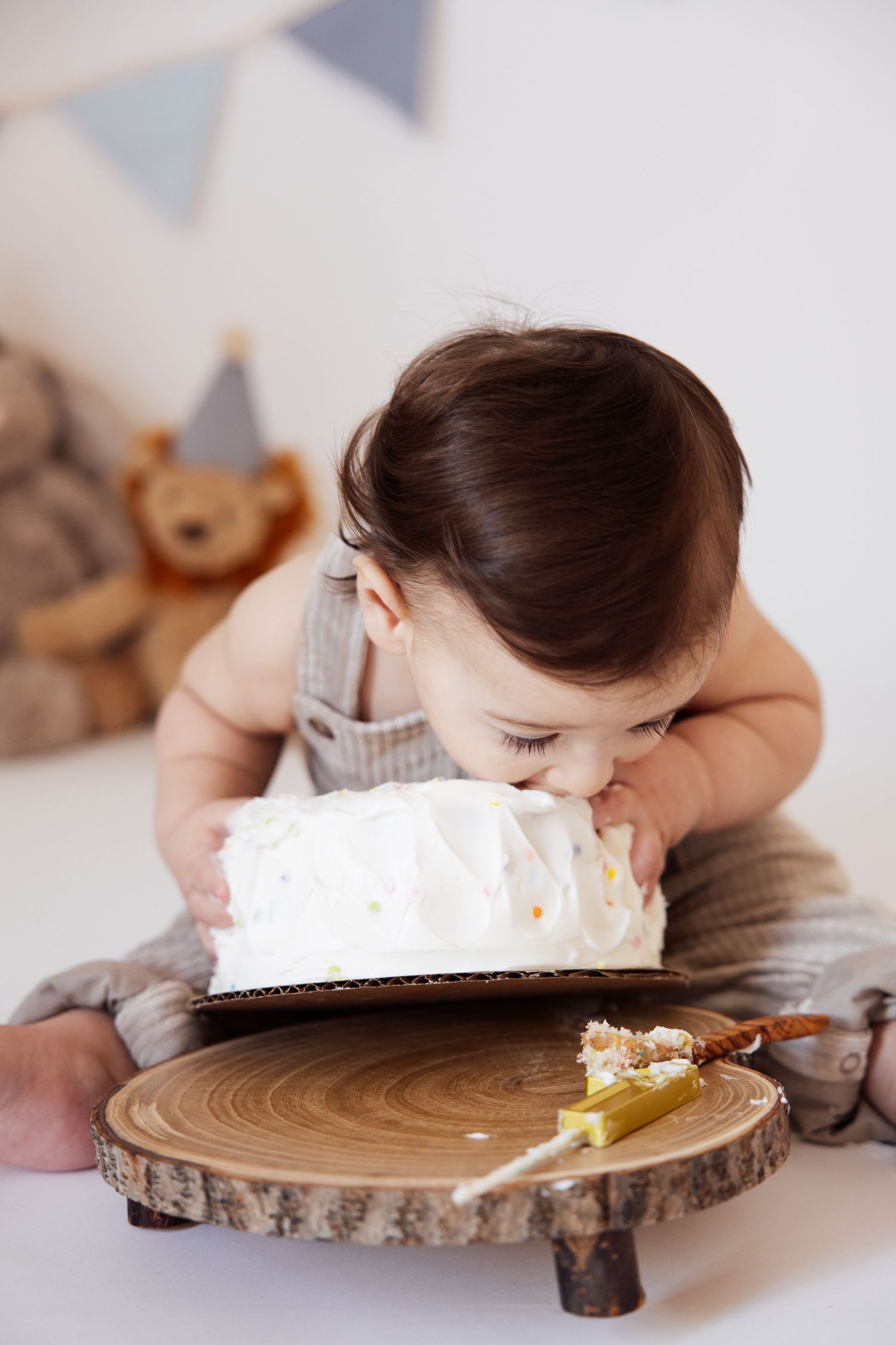 Curious baby in gray overalls leaning into a white frosted smash cake on a rustic wooden cake stand.