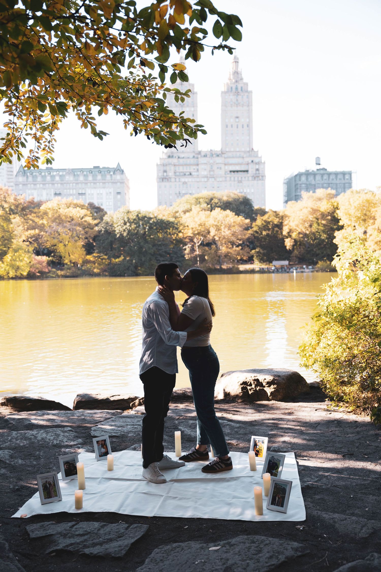 Couple sharing a kiss beside a romantic lakeside proposal setup with candles and photos in Central Park.