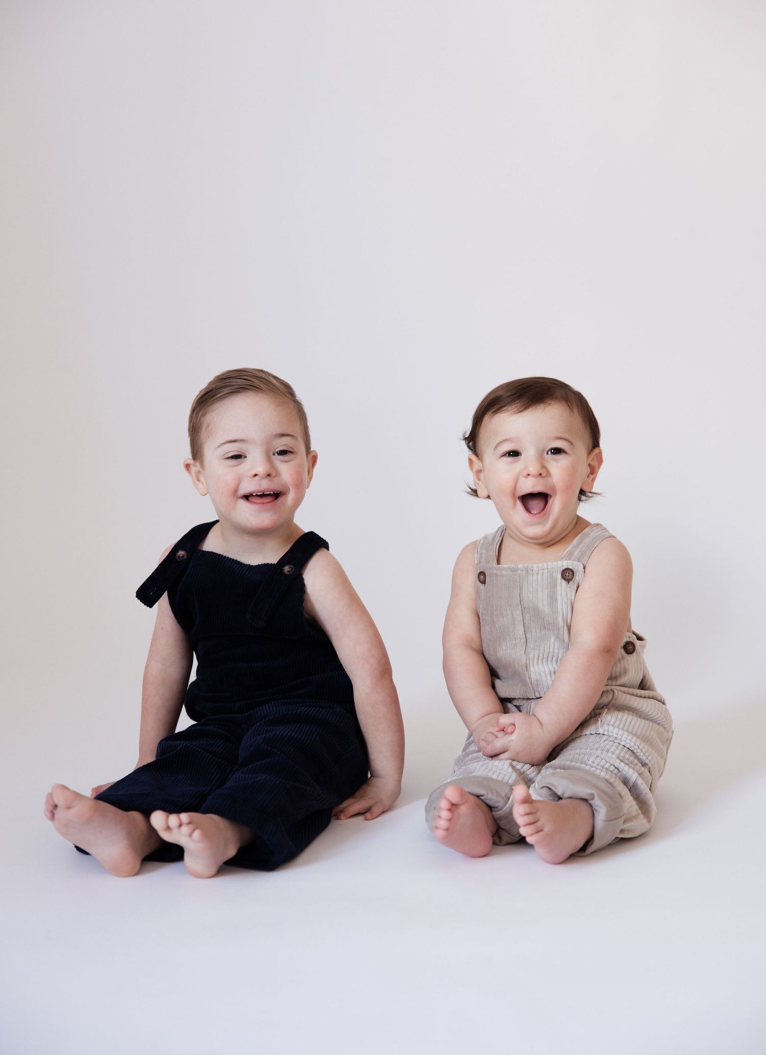 Two joyful babies in navy and tan overalls sitting barefoot together, laughing on a white seamless backdrop.
