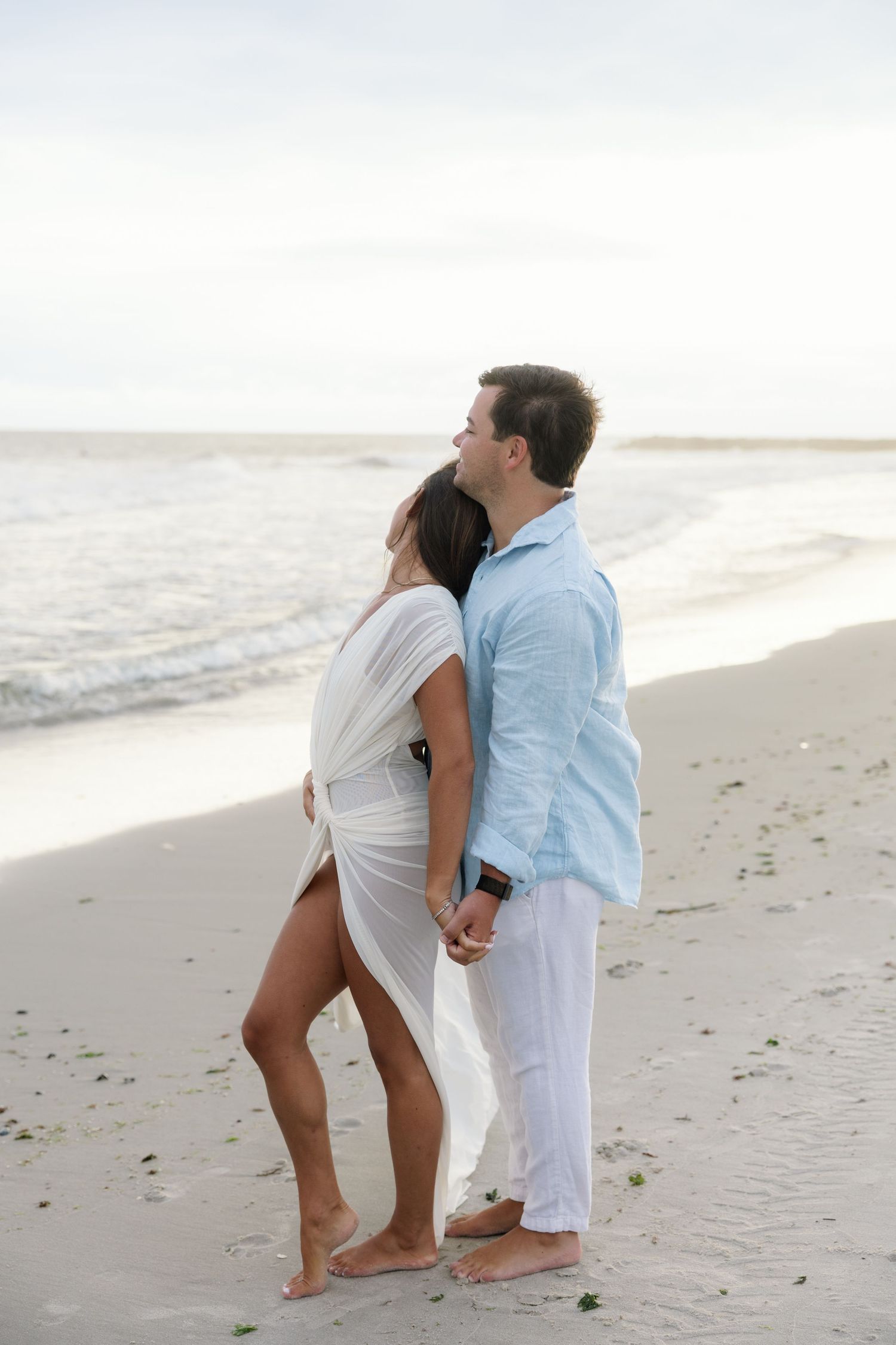 Romantic couple holding hands on a sandy beach, dressed in white and light blue, with ocean waves in the background.