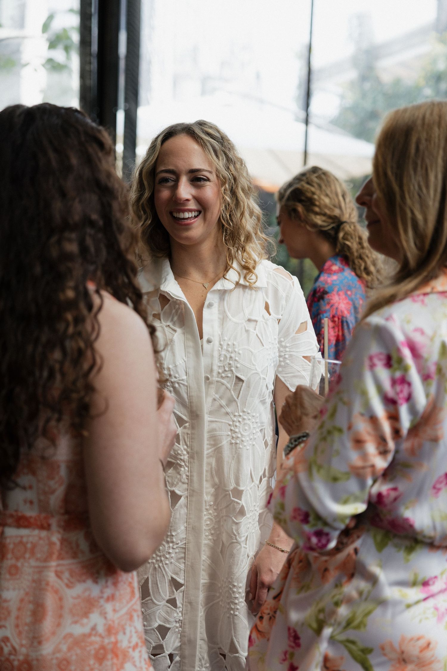 Smiling woman in white lace dress laughing with guests at outdoor bridal shower.