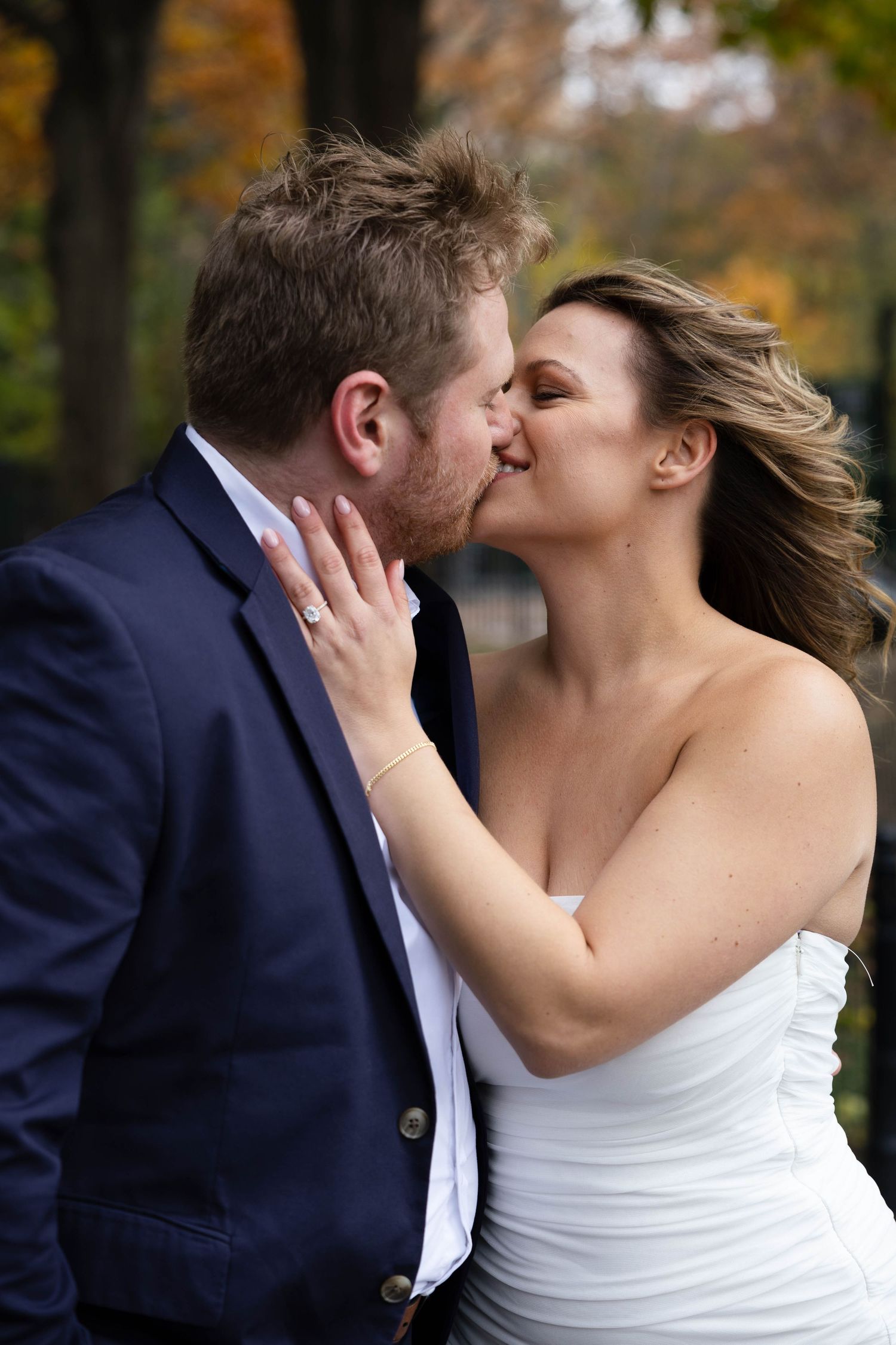 Bride and groom sharing a romantic kiss outdoors with colorful autumn foliage in the background.