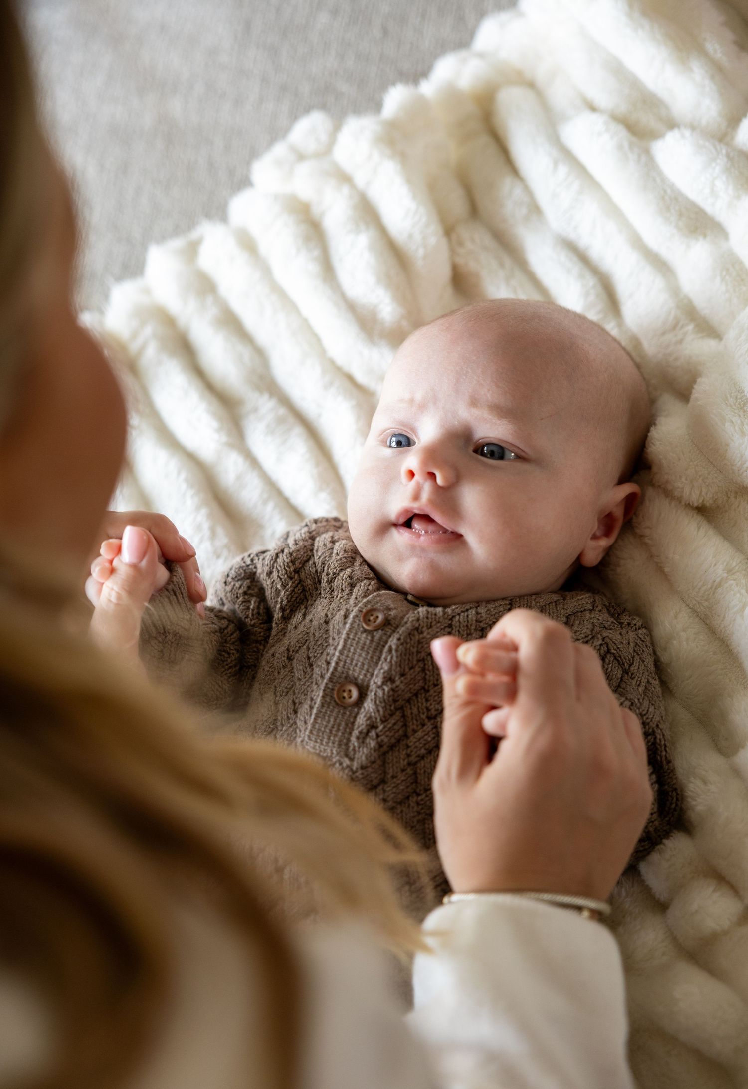 Newborn baby in brown knit cardigan lying on fluffy white blanket, gazing up at mother above.