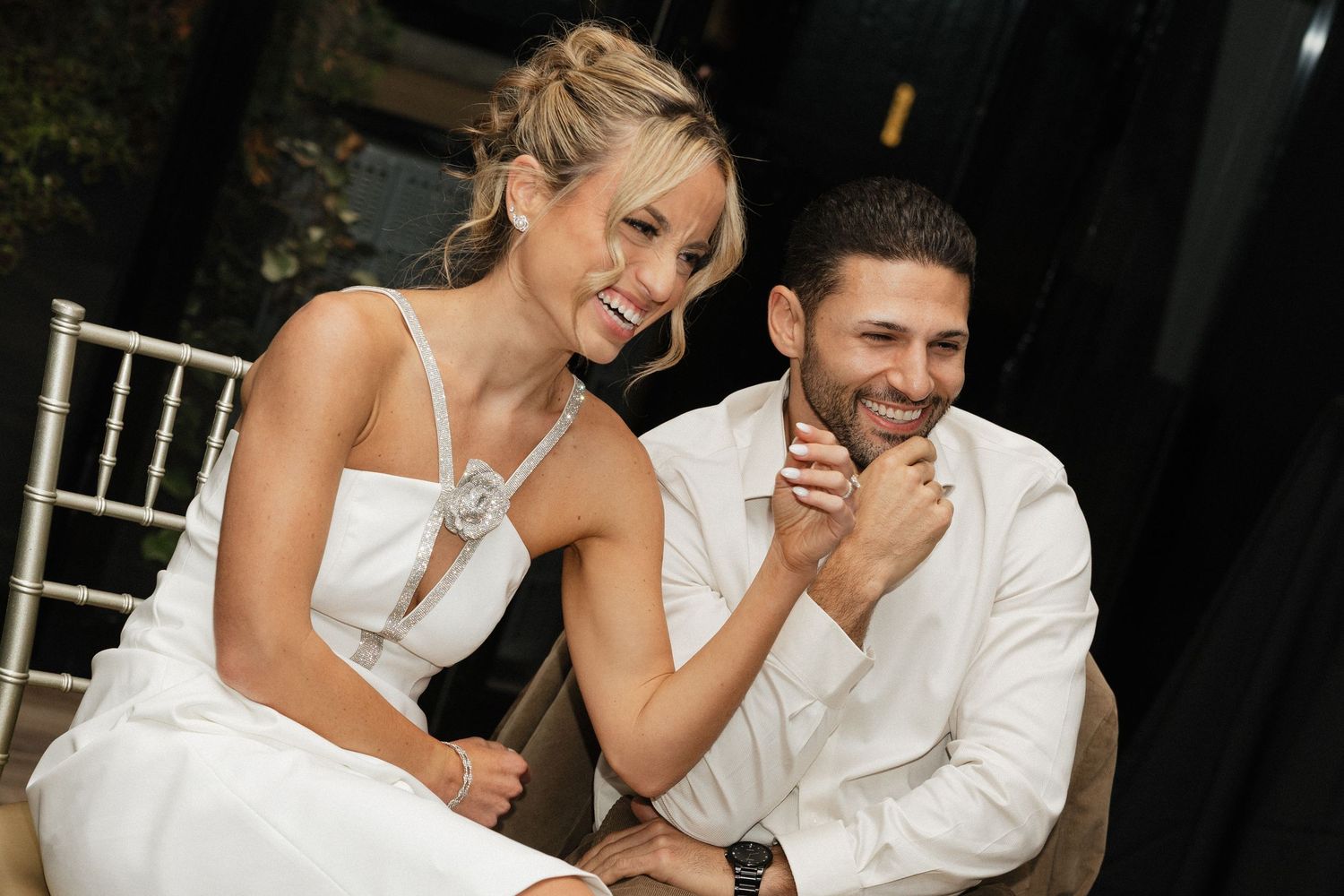 Smiling bride and groom in white outfits seated together at an outdoor wedding reception.