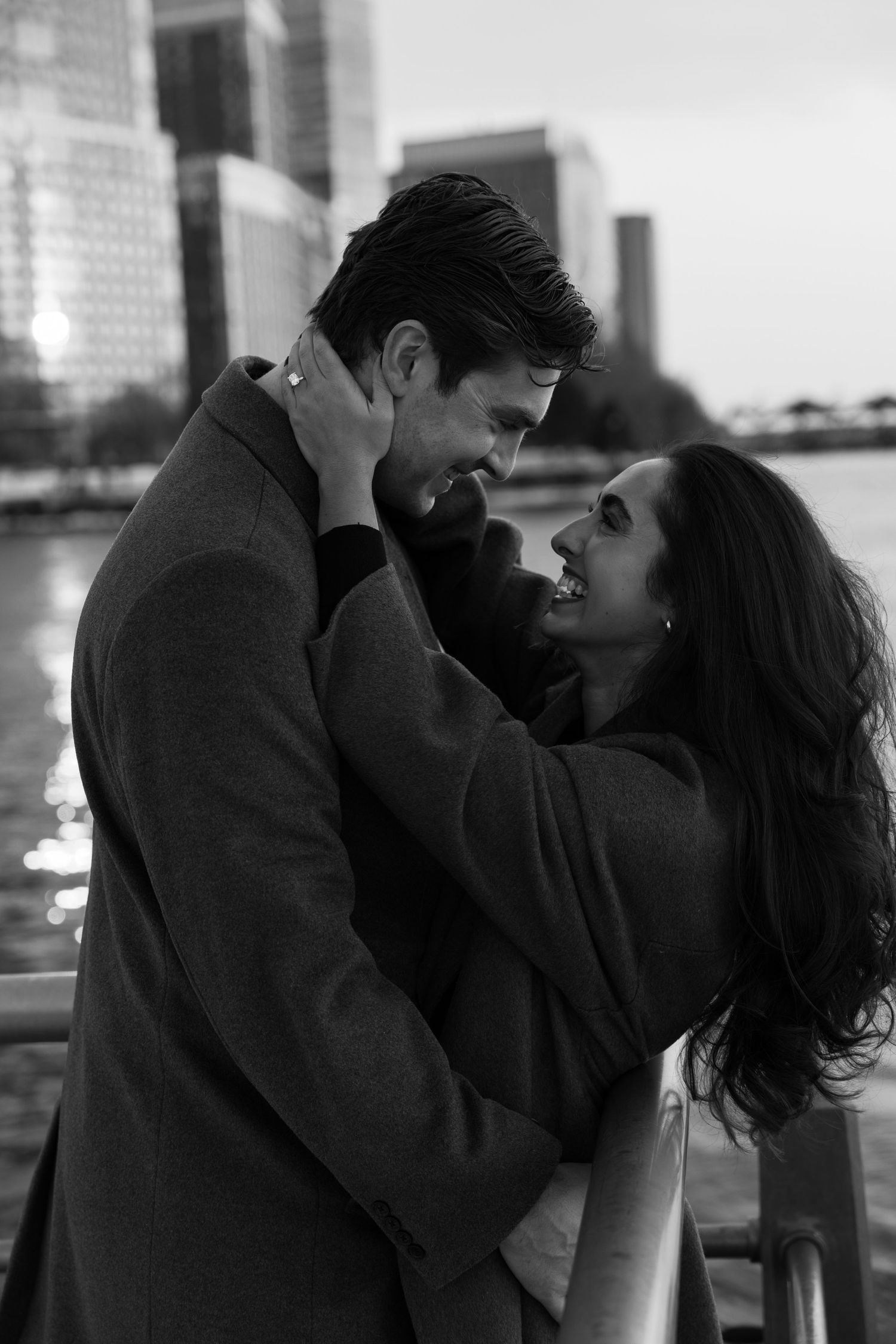 Black and white photo of a romantic couple embracing near a waterfront with city skyline in background.