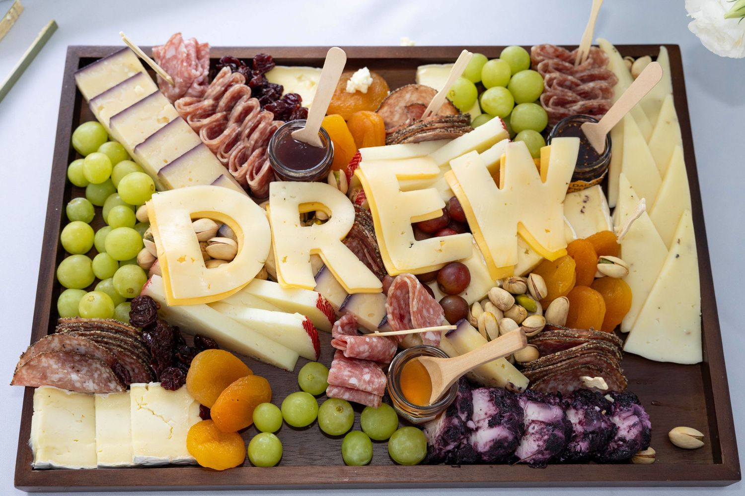 Elegant charcuterie board spelling DREW with cheese letters, grapes, nuts, and dried fruits.