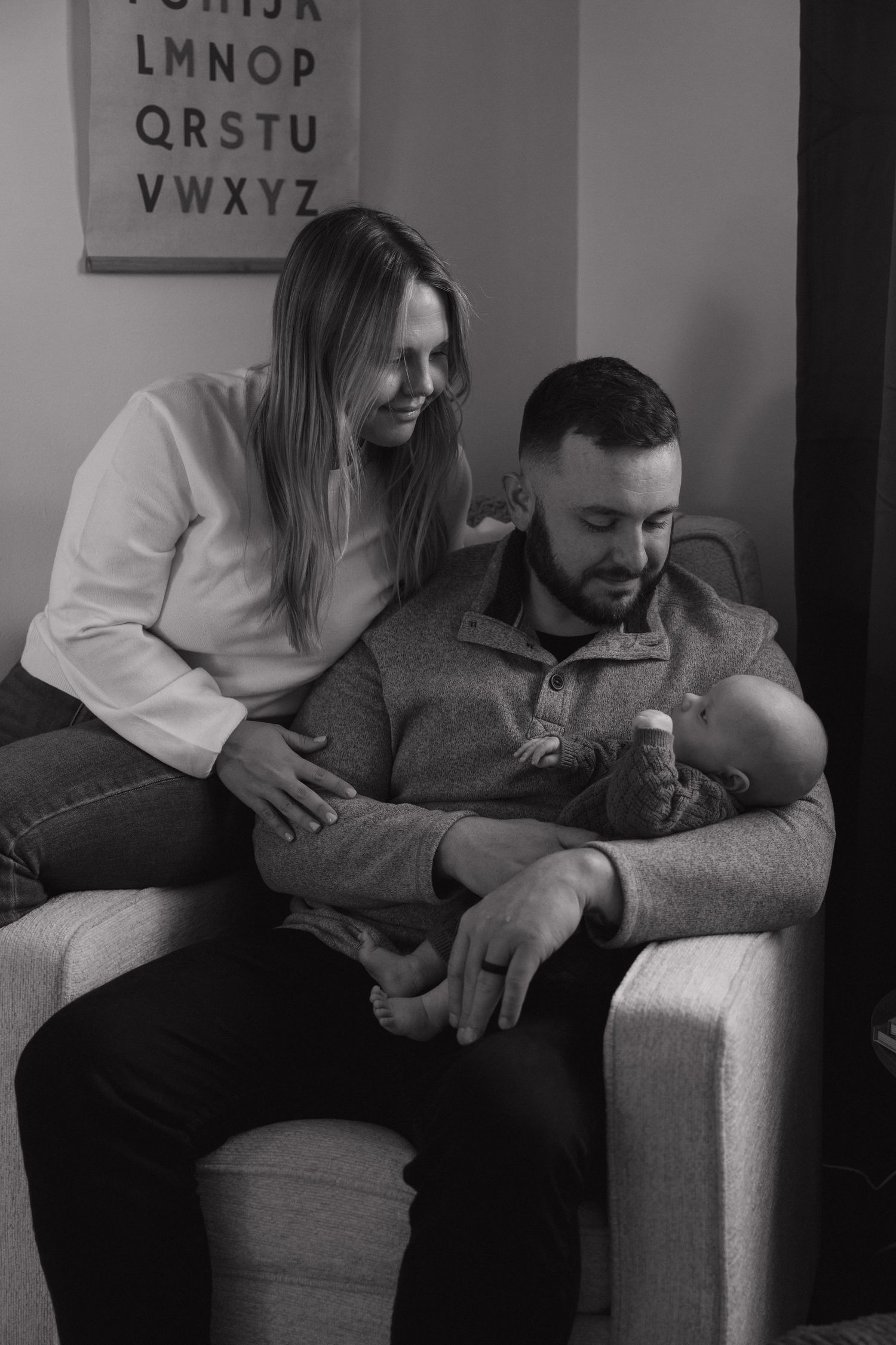 Black and white photo of new parents sitting together in nursery chair, lovingly holding their newborn.