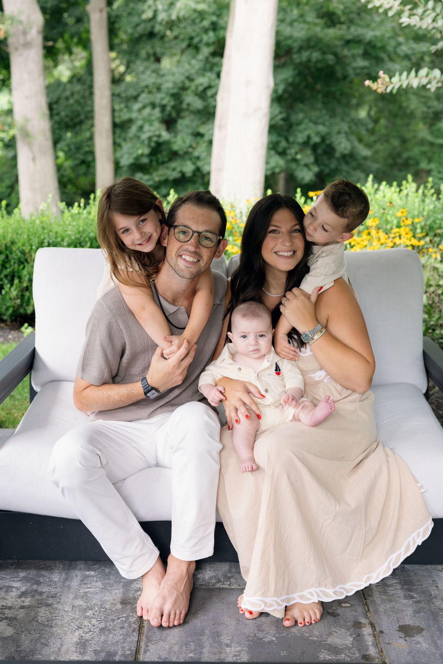 Happy family of five posing outdoors on a white sofa, parents with three young children in a lush garden setting.